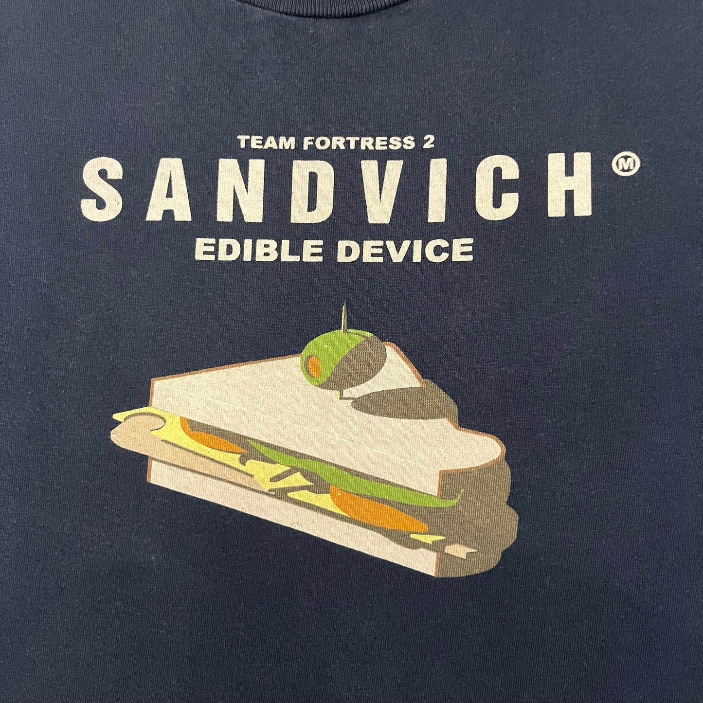 The Team Fortress 2 Sandvich 2010 Tee is an official Valve shirt released in December 2010 and produced during Valve’s in-house merch era. This Navy tee features a Sandvich Edible Device (Heavy secondary weapon) on the front, Sandvich blueprint graphic on the back and 'New Meat!' on the sleeve. A super rare collectible, it represents one of the original Valve Store releases from over a decade ago.
