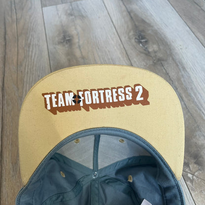 Team Fortress 2 Scout BLU LootCrate Hat is a blue and beige/yellow cap released with the 2016 LootCrate. The blue crown features an embroidered BLU Team Scout Hermes Boot logo, while the beige/yellow visor includes a BLU Team sticker and a large Team Fortress 2 logo on the underside. The hat combines official game branding with collectible LootCrate exclusivity.
