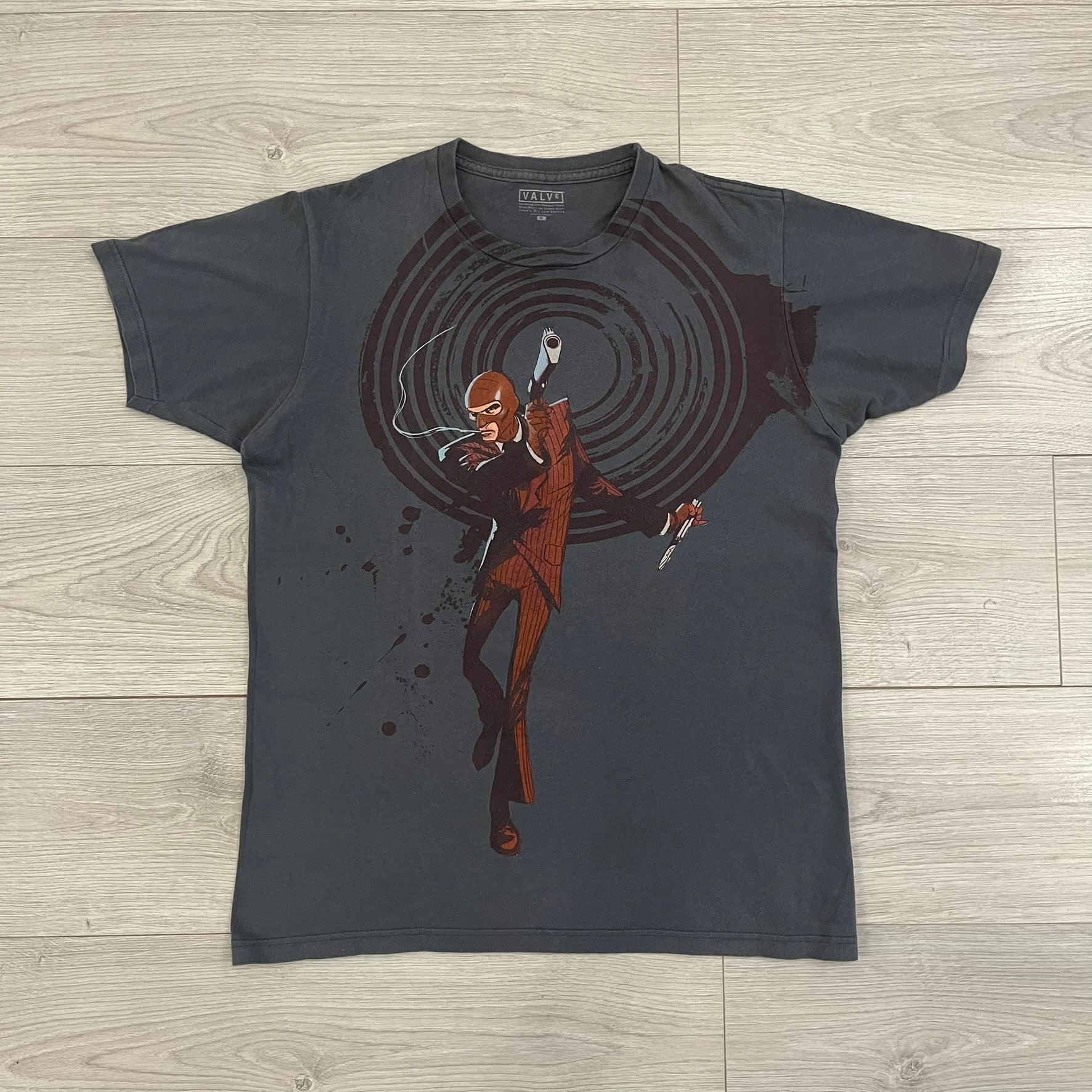 The Team Fortress 2 Spy 2009 Tee is an official Valve shirt from the Sniper vs. Spy update, produced during Valve’s in-house merch era before they switched to a different shirt company. This charcoal tee features a red Spy character holding a butterfly knife and gun, set against a dark red target graphic with dripping effects. A super rare collectible, it represents one of the original Valve Store releases from over a decade ago.
