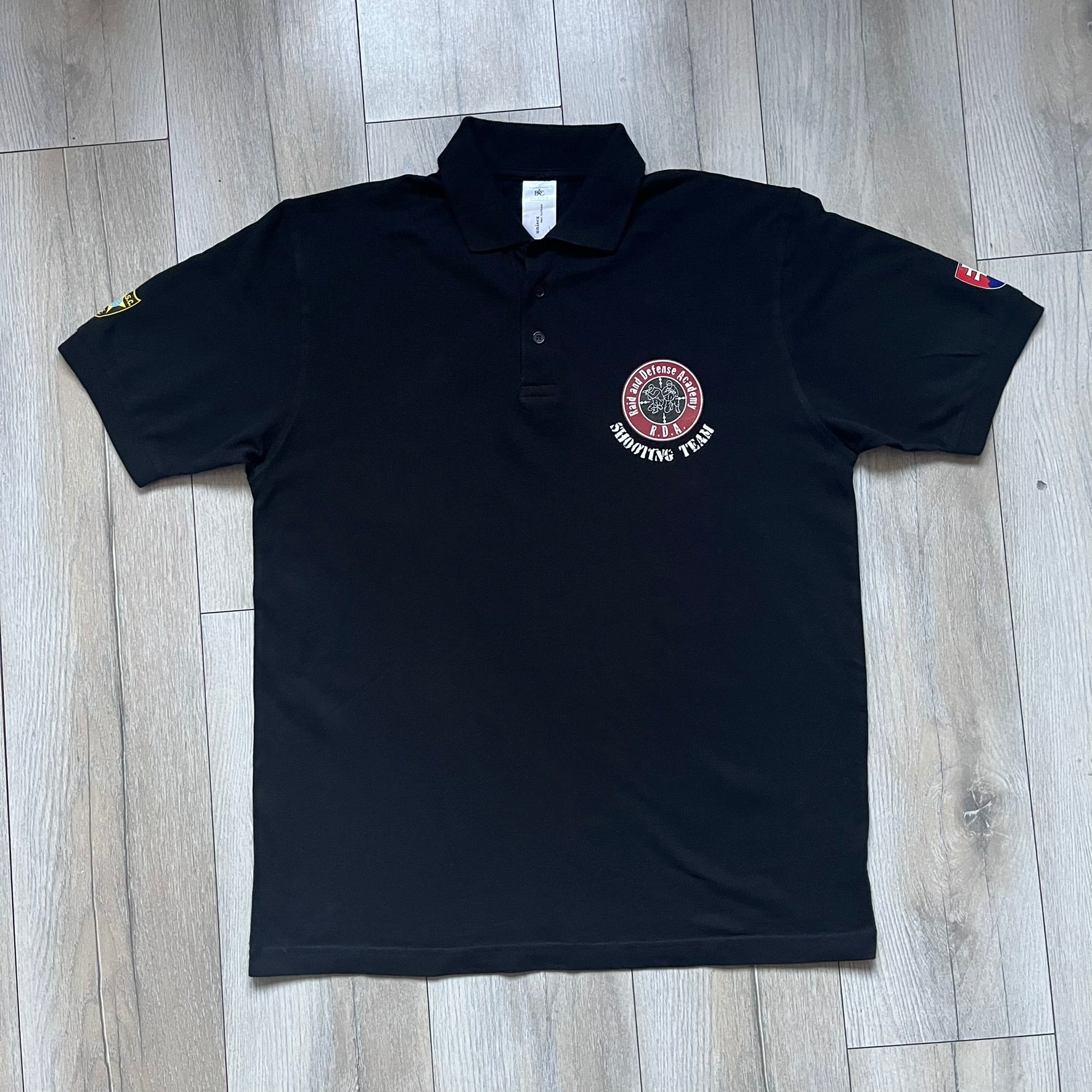 Black polo shirt featuring a large Team Glock logo in white and red printed across the upper back. The chest is marked with the Raid and Defense Academy Shooting Team logo, while the sleeves carry a Slovakia flag badge on one side and an International Practical Shooting Confederation (IPSC) patch on the other. Standard short-sleeve polo construction with a clean, functional finish.
