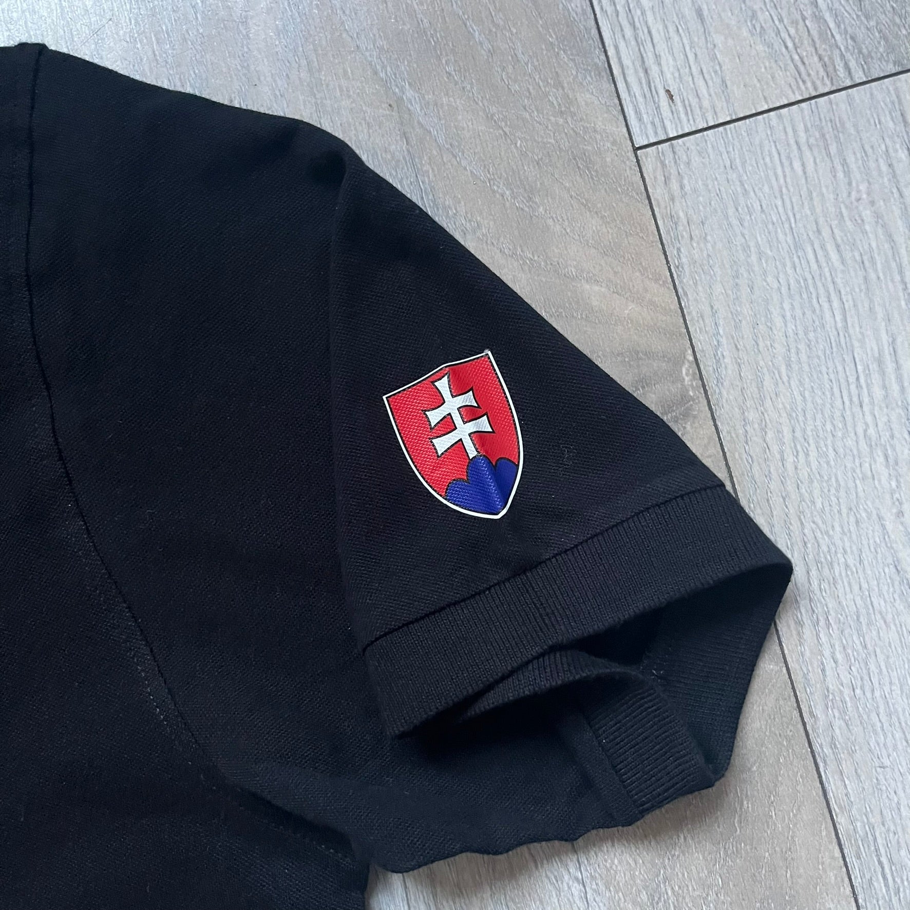 Black polo shirt featuring a large Team Glock logo in white and red printed across the upper back. The chest is marked with the Raid and Defense Academy Shooting Team logo, while the sleeves carry a Slovakia flag badge on one side and an International Practical Shooting Confederation (IPSC) patch on the other. Standard short-sleeve polo construction with a clean, functional finish.