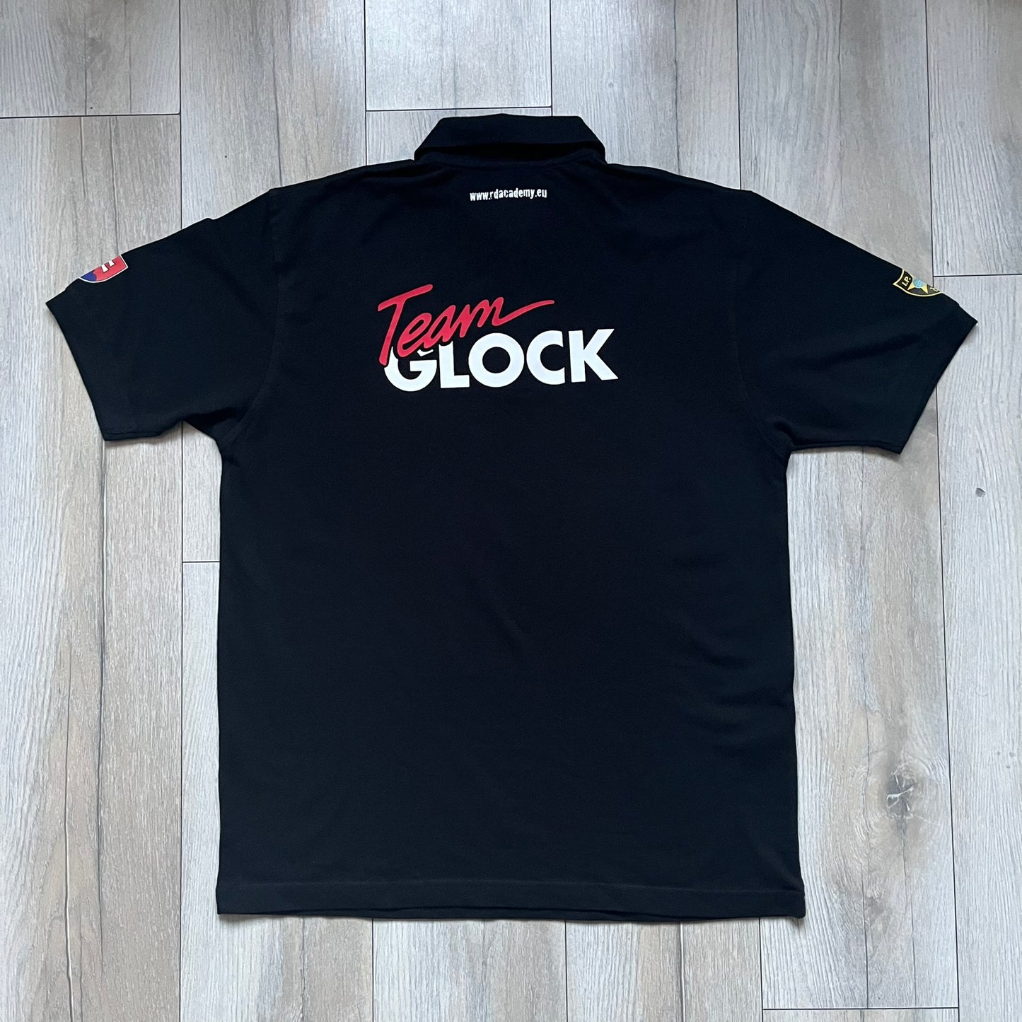 Black polo shirt featuring a large Team Glock logo in white and red printed across the upper back. The chest is marked with the Raid and Defense Academy Shooting Team logo, while the sleeves carry a Slovakia flag badge on one side and an International Practical Shooting Confederation (IPSC) patch on the other. Standard short-sleeve polo construction with a clean, functional finish.