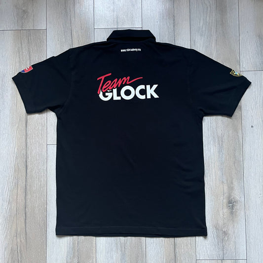 Black polo shirt featuring a large Team Glock logo in white and red printed across the upper back. The chest is marked with the Raid and Defense Academy Shooting Team logo, while the sleeves carry a Slovakia flag badge on one side and an International Practical Shooting Confederation (IPSC) patch on the other. Standard short-sleeve polo construction with a clean, functional finish.