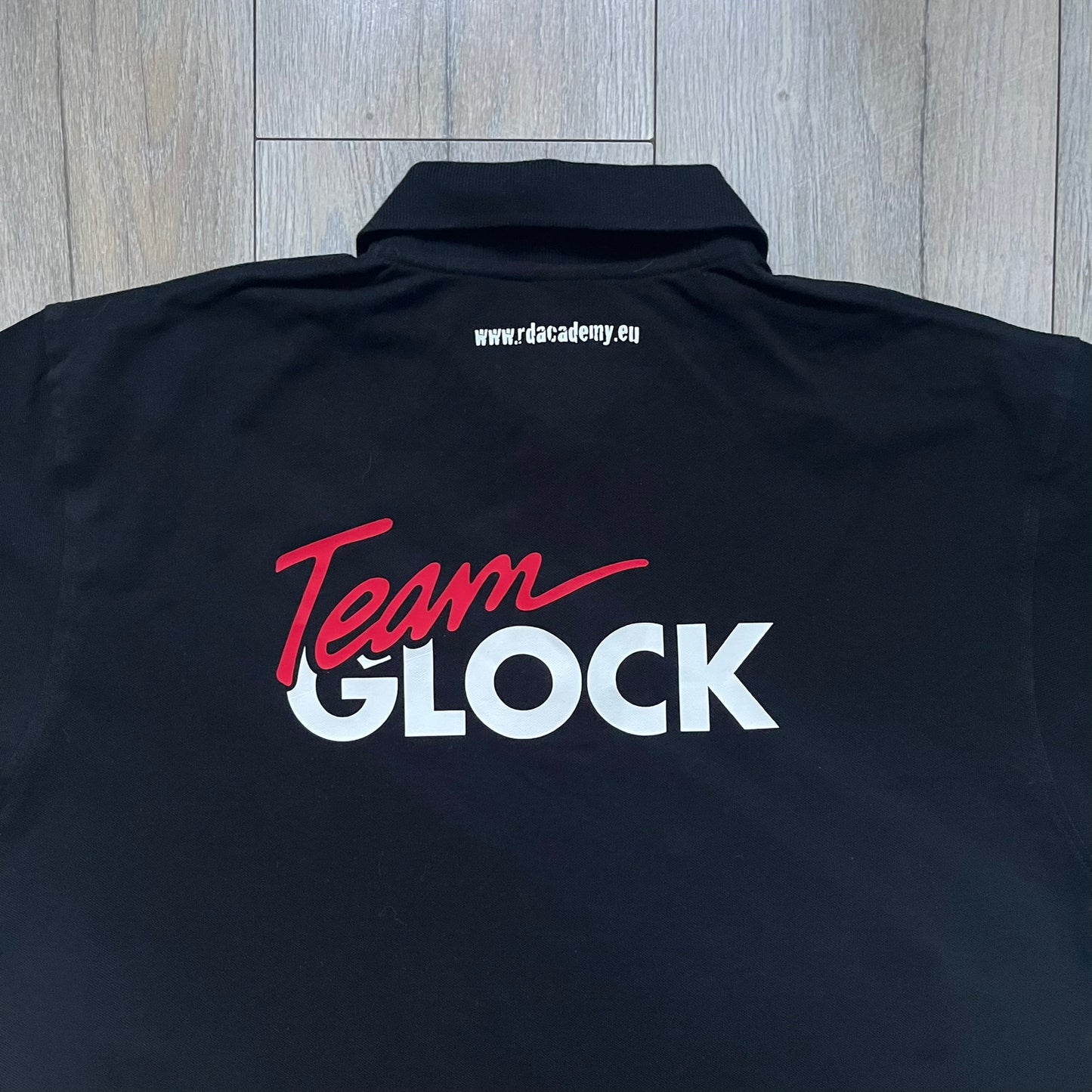 Black polo shirt featuring a large Team Glock logo in white and red printed across the upper back. The chest is marked with the Raid and Defense Academy Shooting Team logo, while the sleeves carry a Slovakia flag badge on one side and an International Practical Shooting Confederation (IPSC) patch on the other. Standard short-sleeve polo construction with a clean, functional finish.