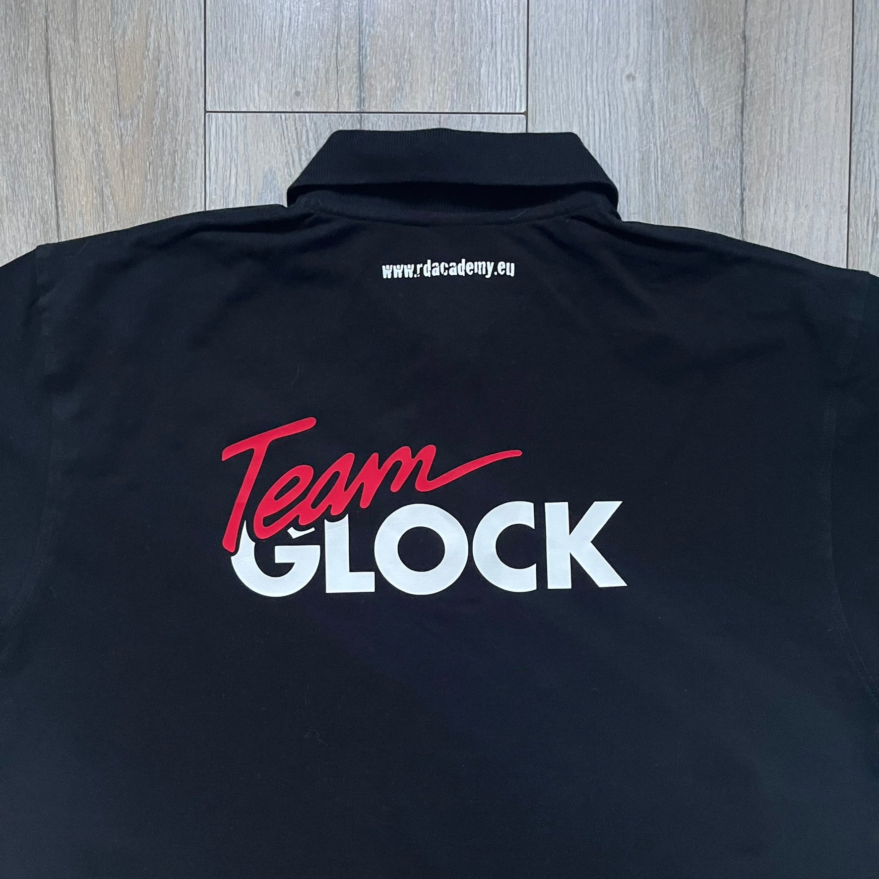 Black polo shirt featuring a large Team Glock logo in white and red printed across the upper back. The chest is marked with the Raid and Defense Academy Shooting Team logo, while the sleeves carry a Slovakia flag badge on one side and an International Practical Shooting Confederation (IPSC) patch on the other. Standard short-sleeve polo construction with a clean, functional finish.