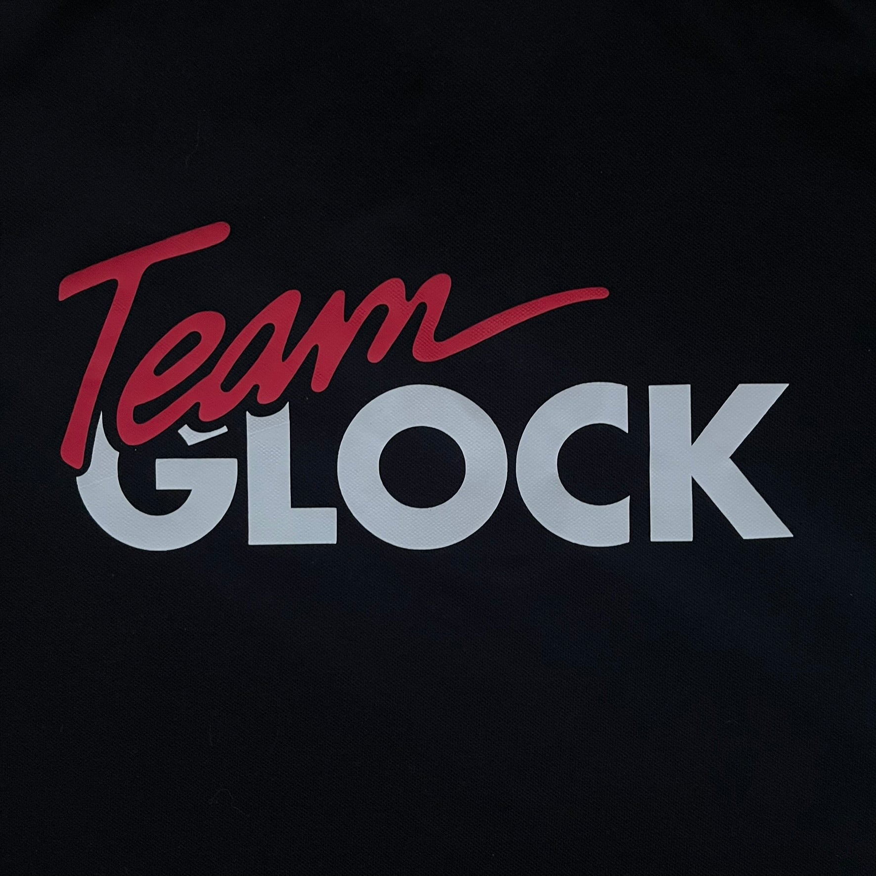 Black polo shirt featuring a large Team Glock logo in white and red printed across the upper back. The chest is marked with the Raid and Defense Academy Shooting Team logo, while the sleeves carry a Slovakia flag badge on one side and an International Practical Shooting Confederation (IPSC) patch on the other. Standard short-sleeve polo construction with a clean, functional finish.