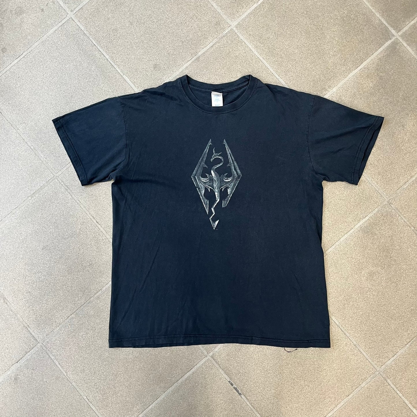 The Elder Scrolls V: Skyrim 2011 Promo Tee features a black design with the Skyrim dragon logo printed on the front chest. The upper back has the full “The Elder Scrolls V: Skyrim” text with Bethesda branding. Made on a Gildan blank.
