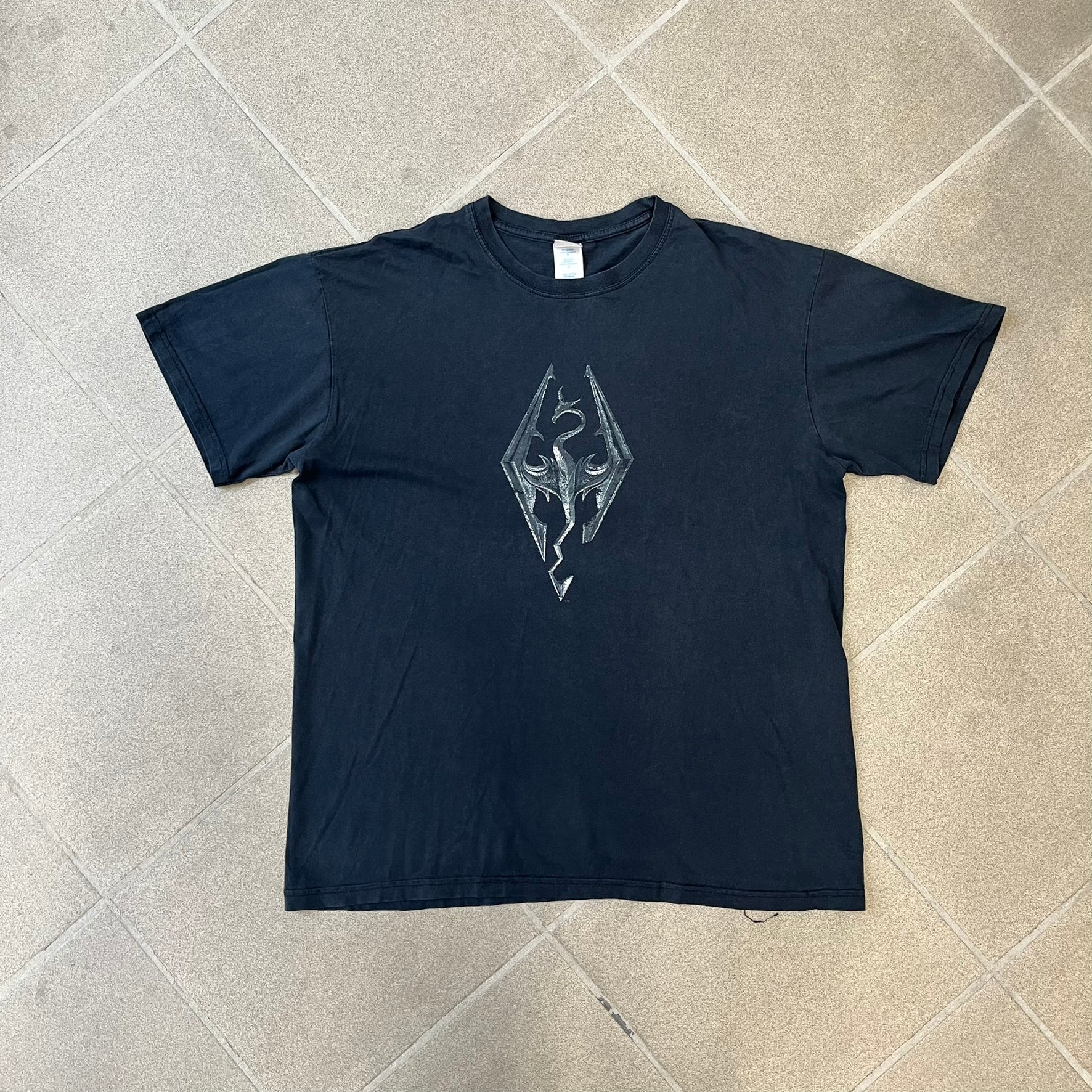 The Elder Scrolls V: Skyrim 2011 Promo Tee features a black design with the Skyrim dragon logo printed on the front chest. The upper back has the full “The Elder Scrolls V: Skyrim” text with Bethesda branding. Made on a Gildan blank.
