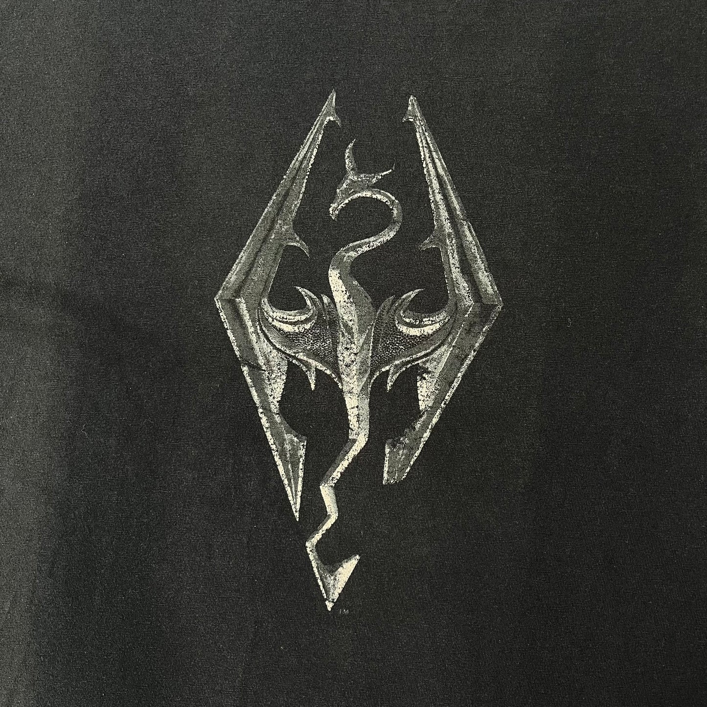 The Elder Scrolls V: Skyrim 2011 Promo Tee features a black design with the Skyrim dragon logo printed on the front chest. The upper back has the full “The Elder Scrolls V: Skyrim” text with Bethesda branding. Made on a Gildan blank.
