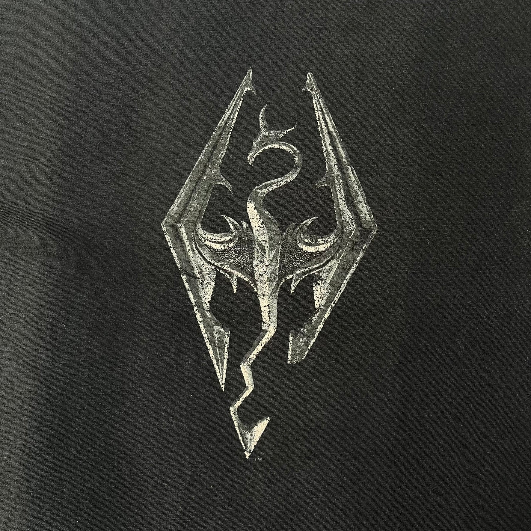 The Elder Scrolls V: Skyrim 2011 Promo Tee features a black design with the Skyrim dragon logo printed on the front chest. The upper back has the full “The Elder Scrolls V: Skyrim” text with Bethesda branding. Made on a Gildan blank.
