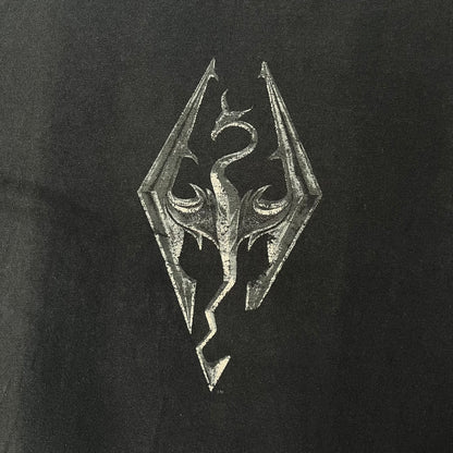 The Elder Scrolls V: Skyrim 2011 Promo Tee features a black design with the Skyrim dragon logo printed on the front chest. The upper back has the full “The Elder Scrolls V: Skyrim” text with Bethesda branding. Made on a Gildan blank.
