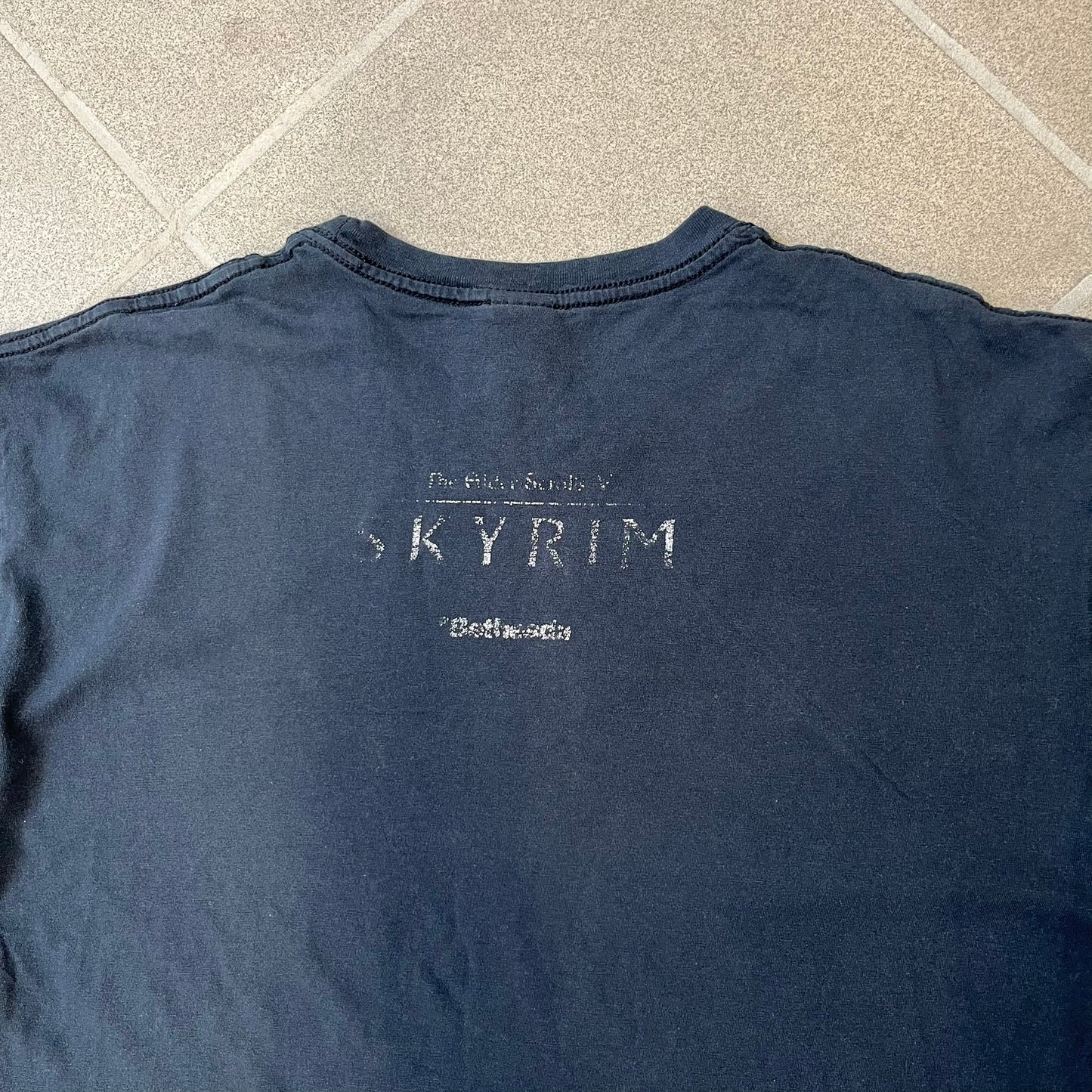 The Elder Scrolls V: Skyrim 2011 Promo Tee features a black design with the Skyrim dragon logo printed on the front chest. The upper back has the full “The Elder Scrolls V: Skyrim” text with Bethesda branding. Made on a Gildan blank.
