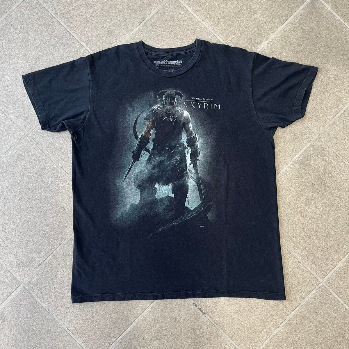 The Elder Scrolls V: Skyrim 2012 Dragonborn Tee features a black 100% cotton construction with the Dragonborn standing on top of a mountain, holding weapons and wearing a horned helmet. The Skyrim logo appears beside the character, with small white “@BSW” text underneath. Finished with a Bethesda licensed neck tag.
