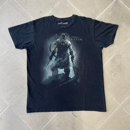 The Elder Scrolls V: Skyrim 2012 Dragonborn Tee features a black 100% cotton construction with the Dragonborn standing on top of a mountain, holding weapons and wearing a horned helmet. The Skyrim logo appears beside the character, with small white “@BSW” text underneath. Finished with a Bethesda licensed neck tag.
