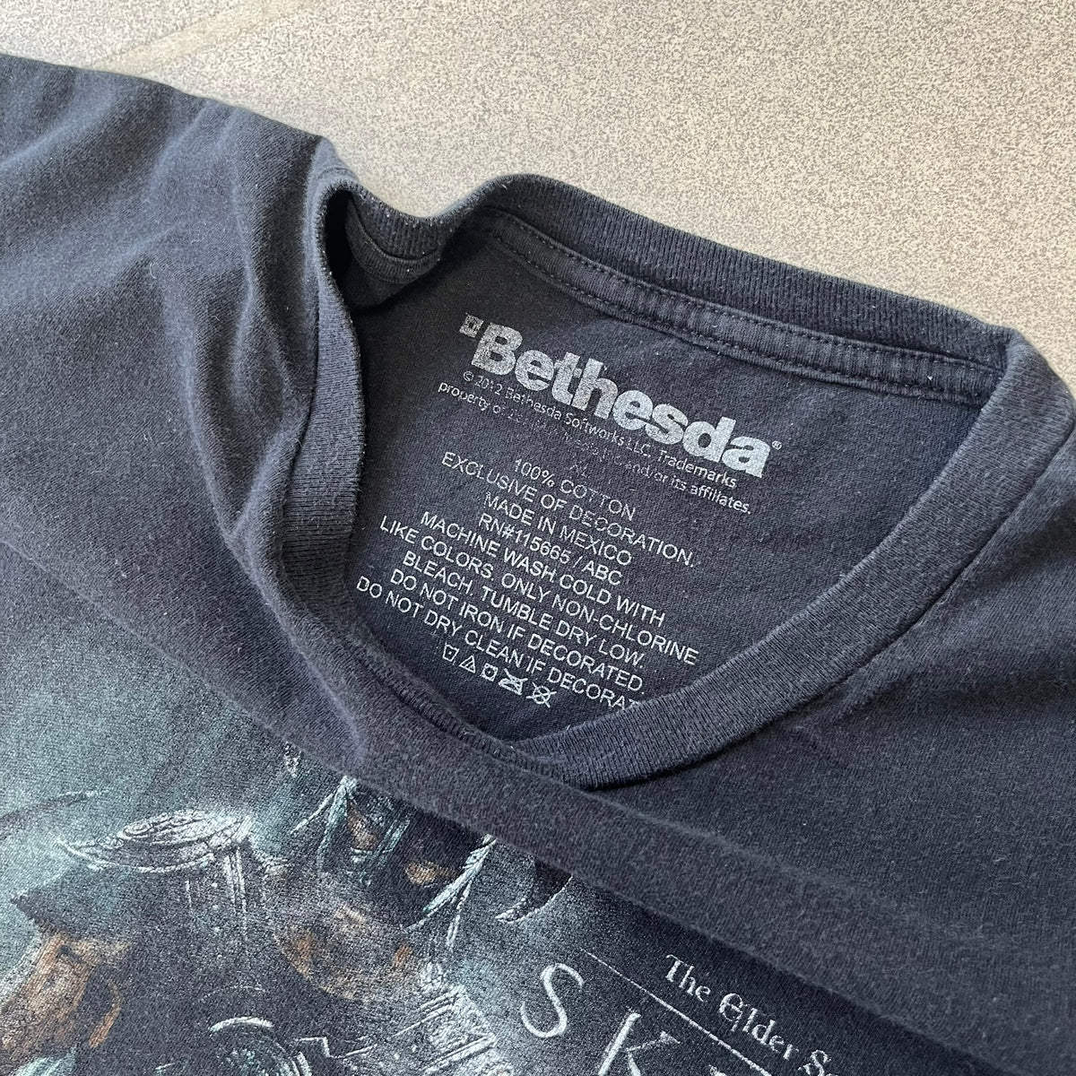 The Elder Scrolls V: Skyrim 2012 Dragonborn Tee features a black 100% cotton construction with the Dragonborn standing on top of a mountain, holding weapons and wearing a horned helmet. The Skyrim logo appears beside the character, with small white “@BSW” text underneath. Finished with a Bethesda licensed neck tag.
