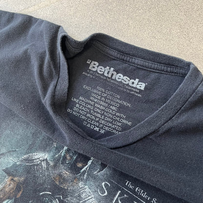 The Elder Scrolls V: Skyrim 2012 Dragonborn Tee features a black 100% cotton construction with the Dragonborn standing on top of a mountain, holding weapons and wearing a horned helmet. The Skyrim logo appears beside the character, with small white “@BSW” text underneath. Finished with a Bethesda licensed neck tag.
