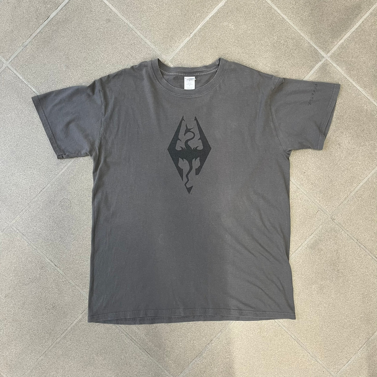 The Elder Scrolls V: Skyrim Premium Edition Tee comes from the 2012 Premium release of the game. Dark gray design with the Skyrim dragon logo printed on the front chest. The upper back has the full “The Elder Scrolls V: Skyrim” text. On the left sleeve there is a text in Dragon language. Made on a Gildan blank.