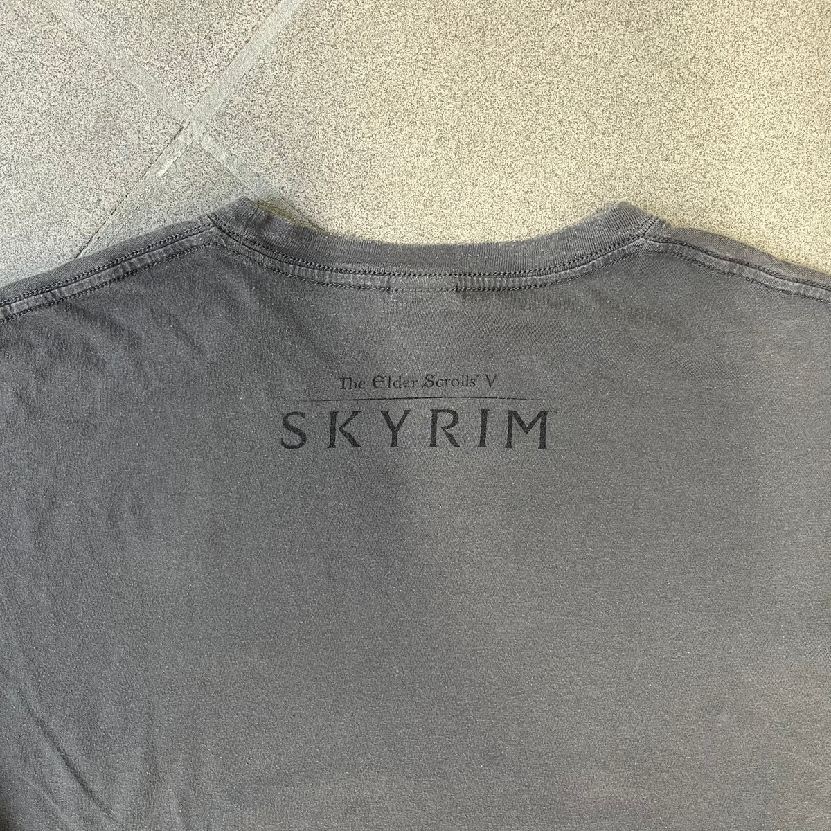 The Elder Scrolls V: Skyrim Premium Edition Tee comes from the 2012 Premium release of the game. Dark gray design with the Skyrim dragon logo printed on the front chest. The upper back has the full “The Elder Scrolls V: Skyrim” text. On the left sleeve there is a text in Dragon language. Made on a Gildan blank.