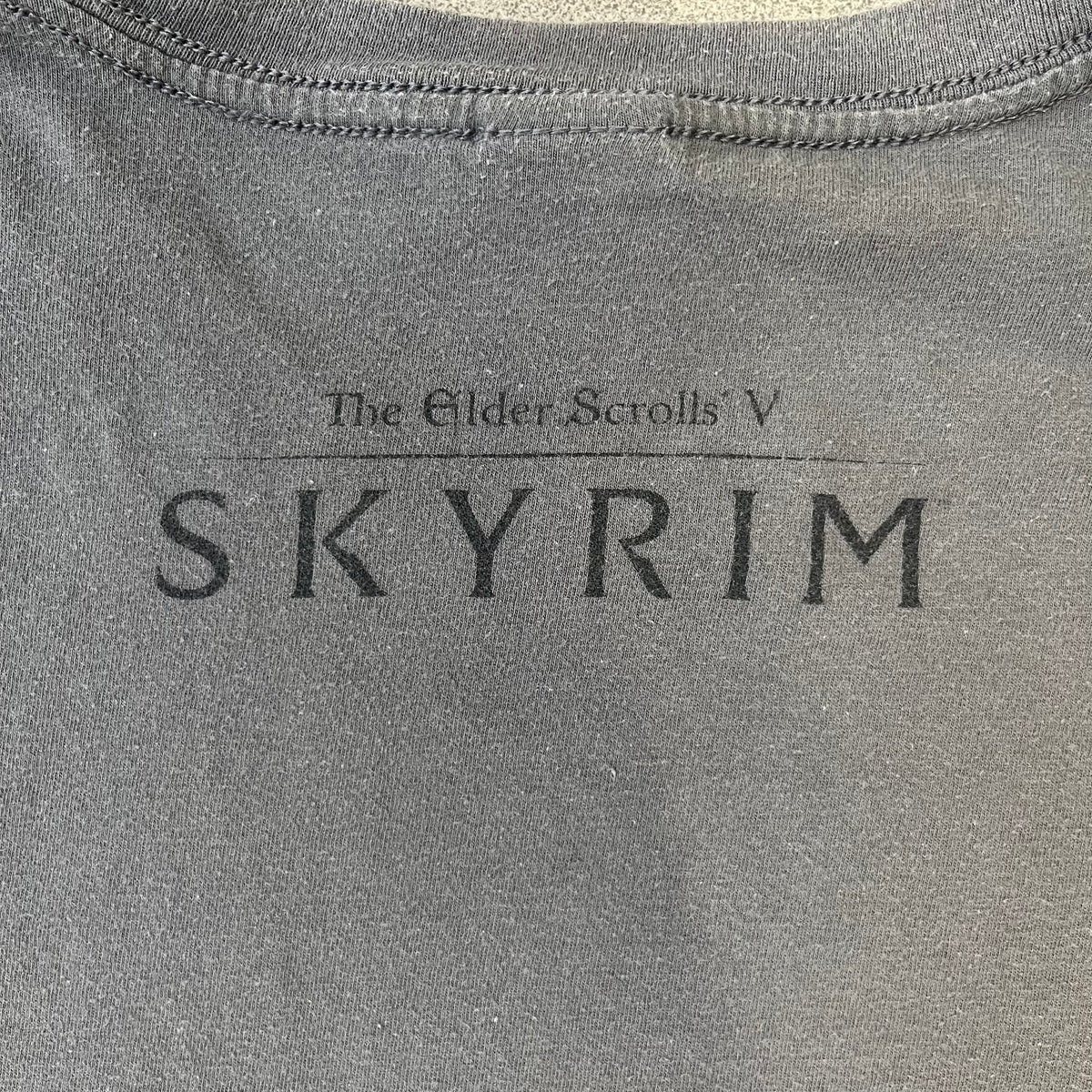 The Elder Scrolls V: Skyrim Premium Edition Tee comes from the 2012 Premium release of the game. Dark gray design with the Skyrim dragon logo printed on the front chest. The upper back has the full “The Elder Scrolls V: Skyrim” text. On the left sleeve there is a text in Dragon language. Made on a Gildan blank.