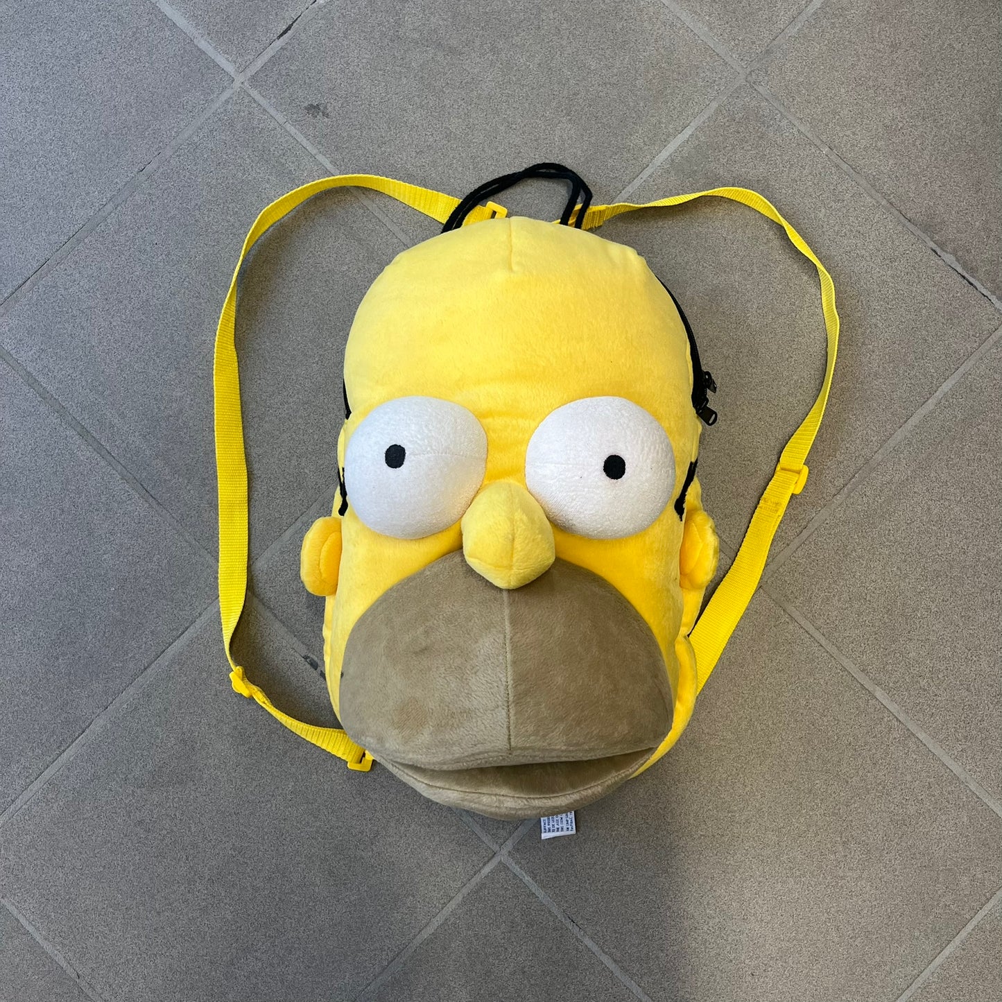 The Simpsons 1990s Homer Simpson Head Backpack is a novelty backpack shaped like a 3D Homer Simpson head. The design is fully yellow with soft sculpted facial features including eyes and nose, a molded mouth, and protruding ears. The hair detail wraps around the back, with the top strands functioning as a hanger. It includes yellow shoulder straps and a single main compartment.
