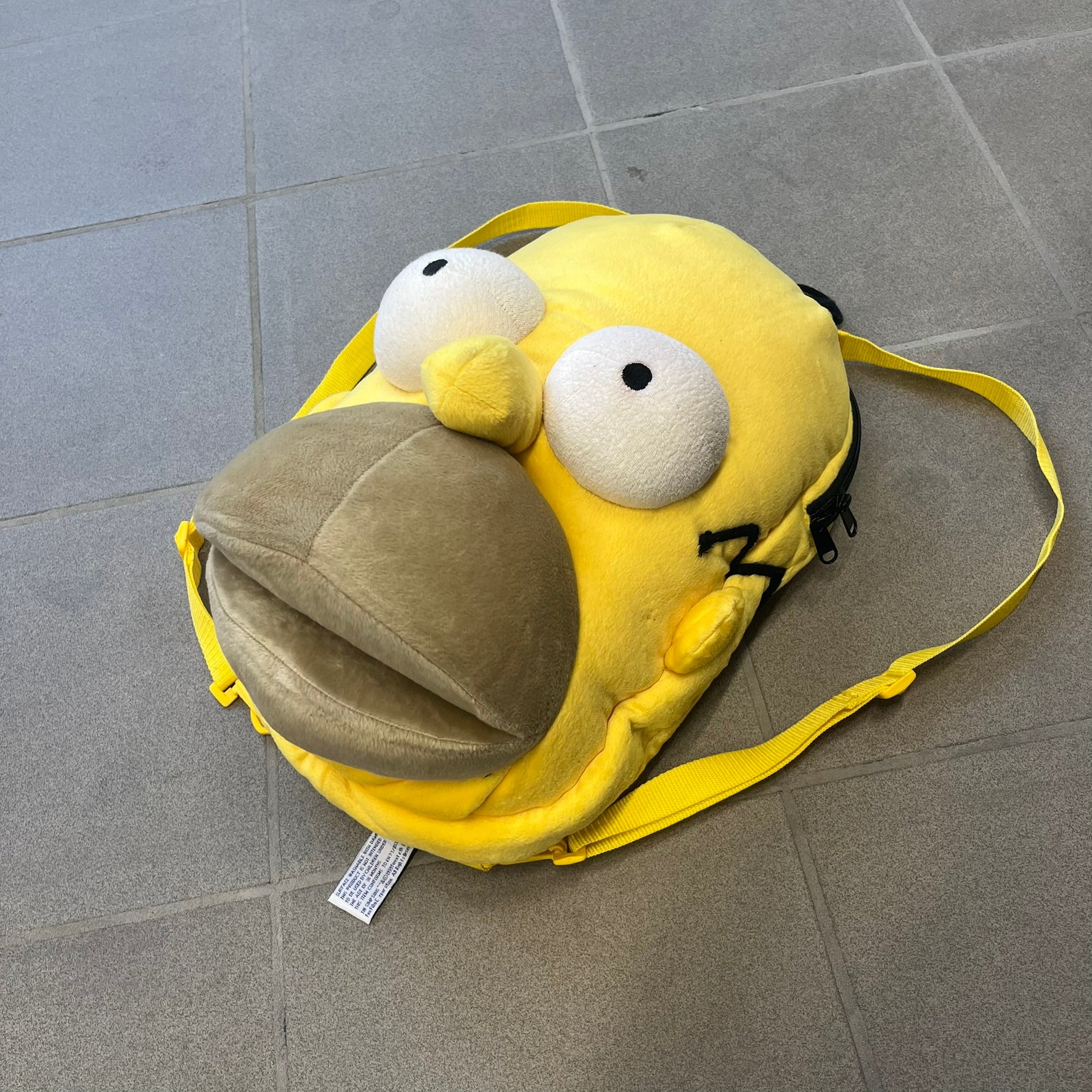 The Simpsons 1990s Homer Simpson Head Backpack is a novelty backpack shaped like a 3D Homer Simpson head. The design is fully yellow with soft sculpted facial features including eyes and nose, a molded mouth, and protruding ears. The hair detail wraps around the back, with the top strands functioning as a hanger. It includes yellow shoulder straps and a single main compartment.