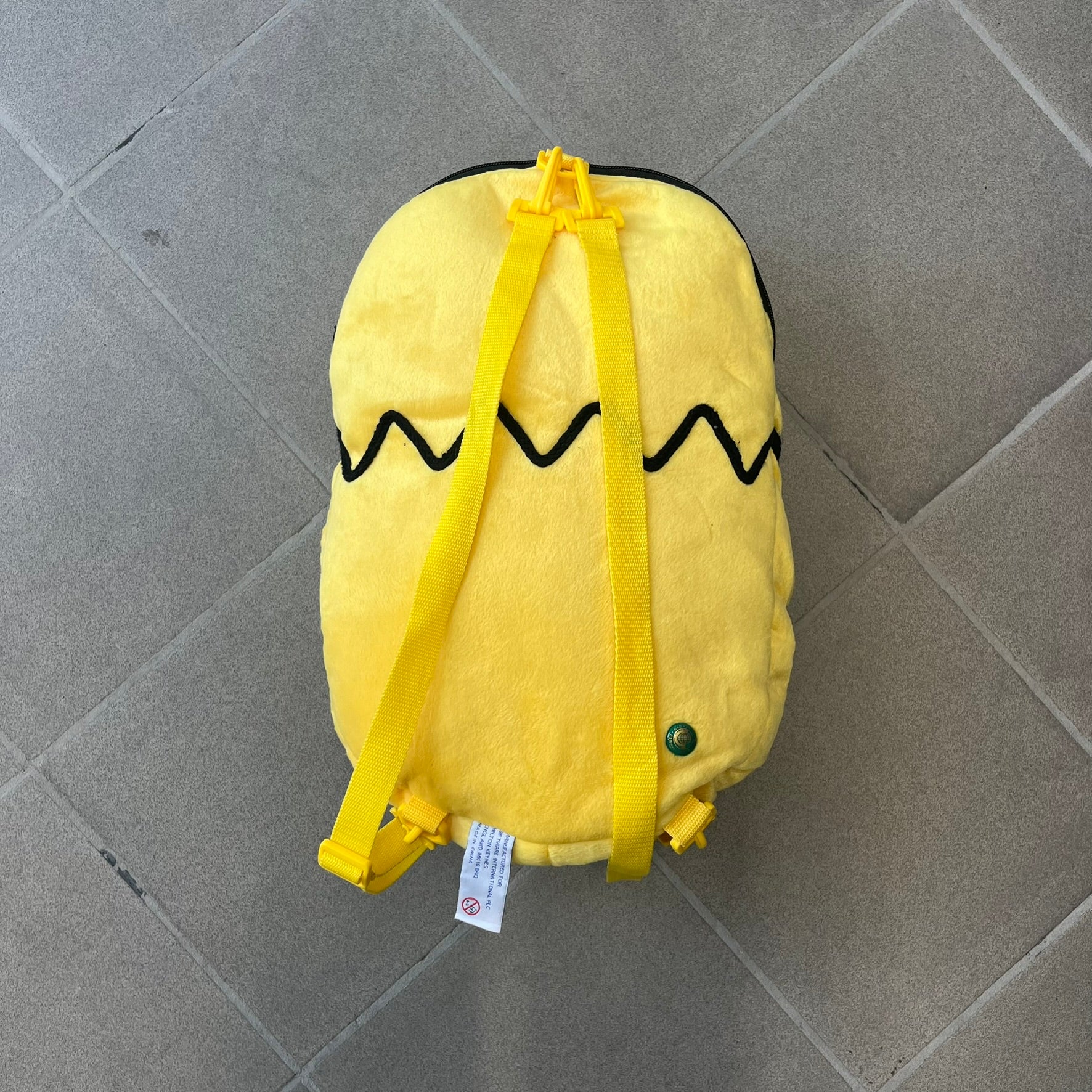 The Simpsons 1990s Homer Simpson Head Backpack is a novelty backpack shaped like a 3D Homer Simpson head. The design is fully yellow with soft sculpted facial features including eyes and nose, a molded mouth, and protruding ears. The hair detail wraps around the back, with the top strands functioning as a hanger. It includes yellow shoulder straps and a single main compartment.