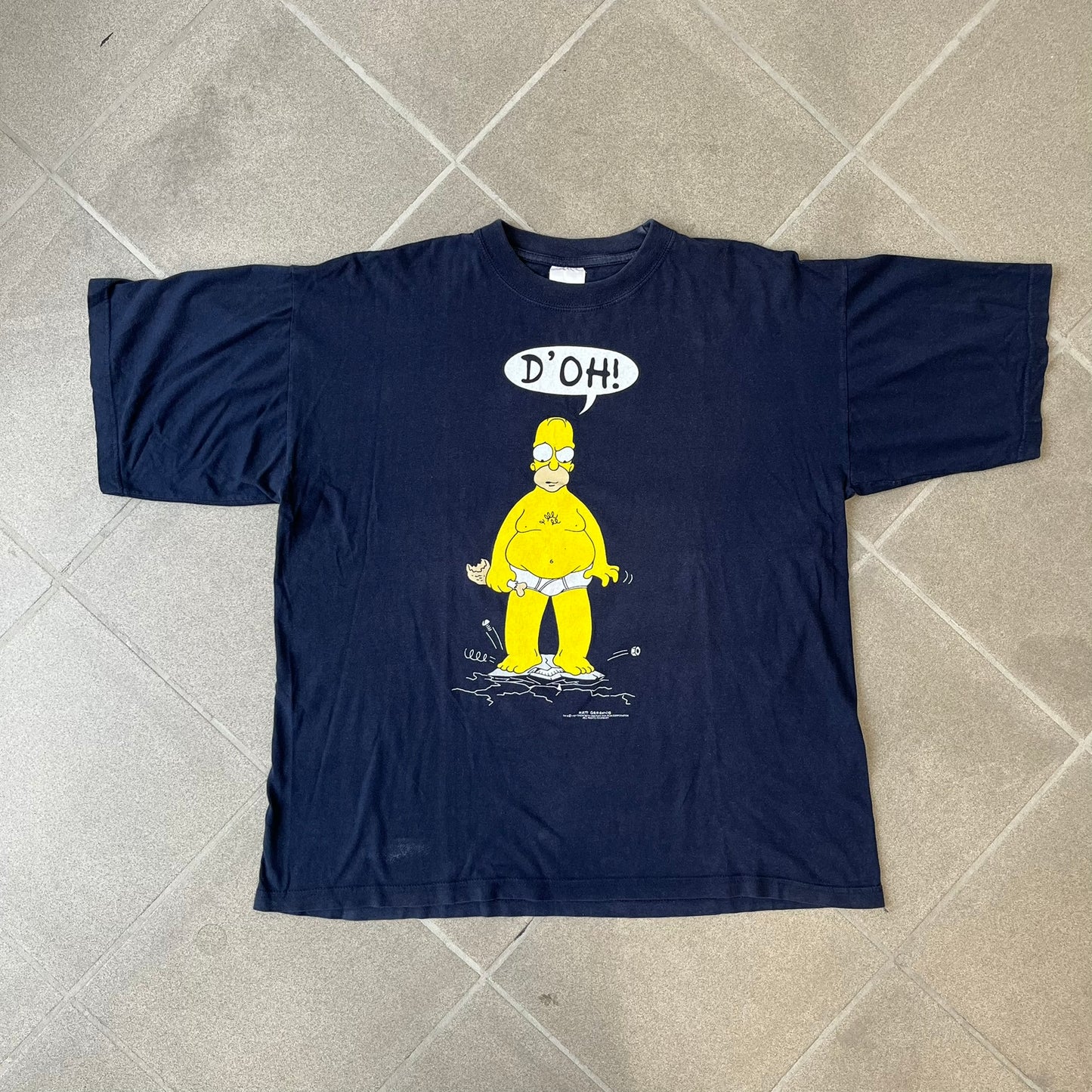 The Simpsons 1997 Homer “D’OH!” Black Tee features a black T-shirt with a front graphic of Homer Simpson standing in his underwear and looking down at a cracked scale while holding a chicken leg. A speech bubble above him reads “D’OH!”, with small Matt Groening 1997 licensing text underneath.
