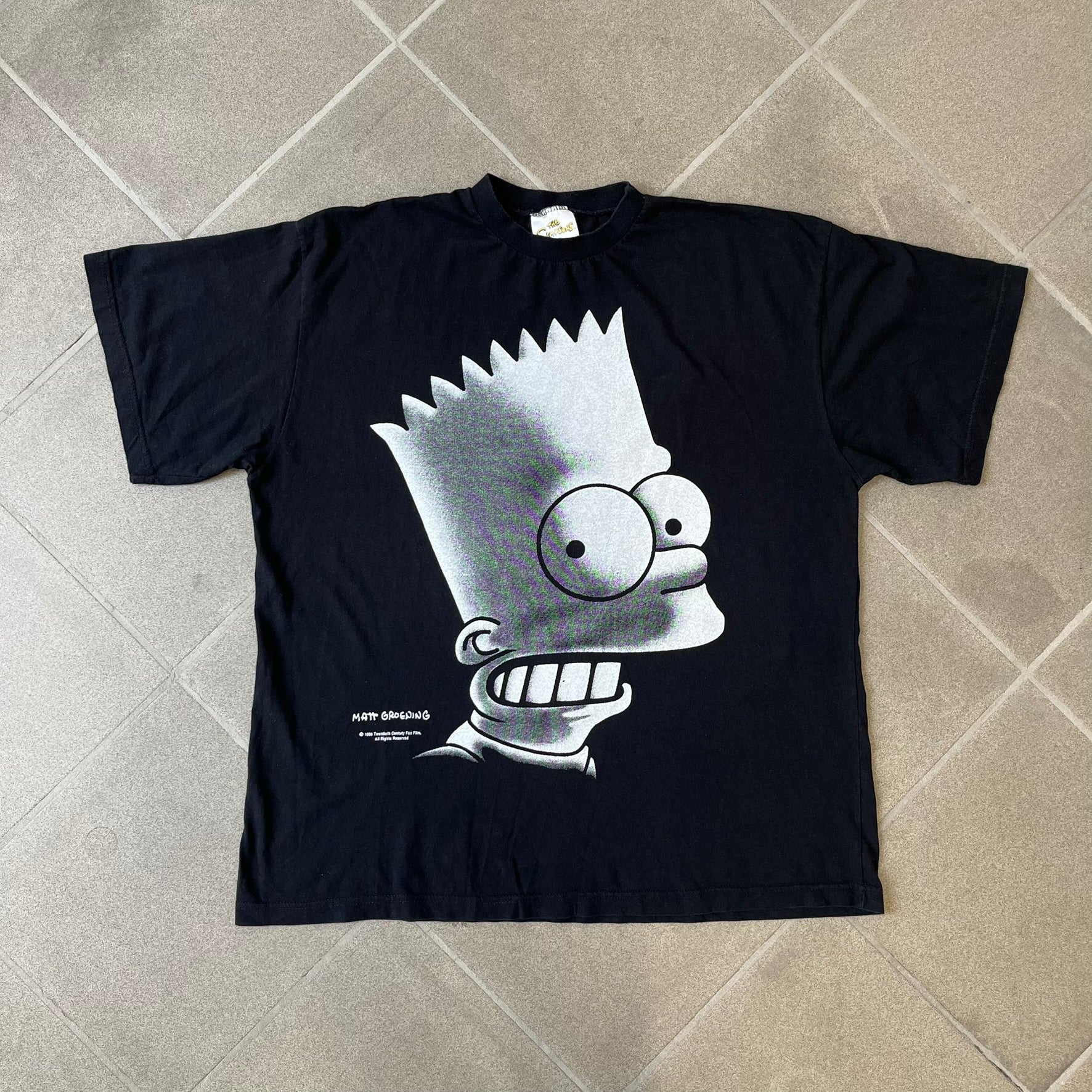 The Simpsons 1999 Bart Face Black Tee features a black T-shirt with a large front graphic of Bart Simpson’s face in black, grey, and white, showing a gritted-teeth expression. Small Matt Groening 1999 licensing text is printed below the graphic.
