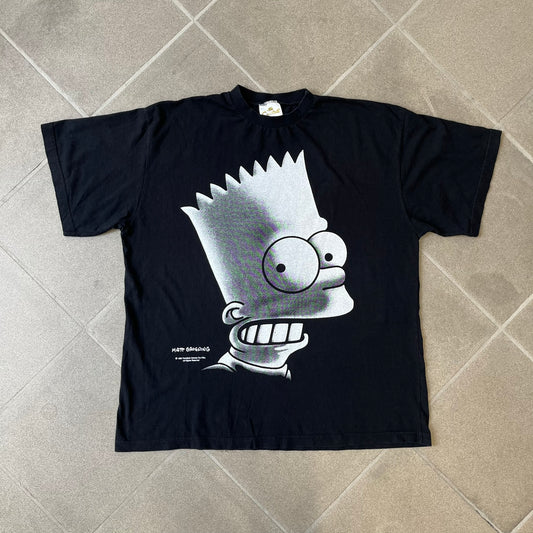 The Simpsons 1999 Bart Face Black Tee features a black T-shirt with a large front graphic of Bart Simpson’s face in black, grey, and white, showing a gritted-teeth expression. Small Matt Groening 1999 licensing text is printed below the graphic.
