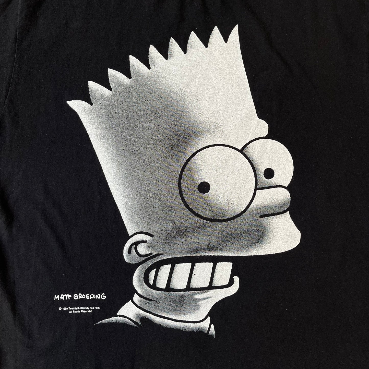 The Simpsons 1999 Bart Face Black Tee features a black T-shirt with a large front graphic of Bart Simpson’s face in black, grey, and white, showing a gritted-teeth expression. Small Matt Groening 1999 licensing text is printed below the graphic.
