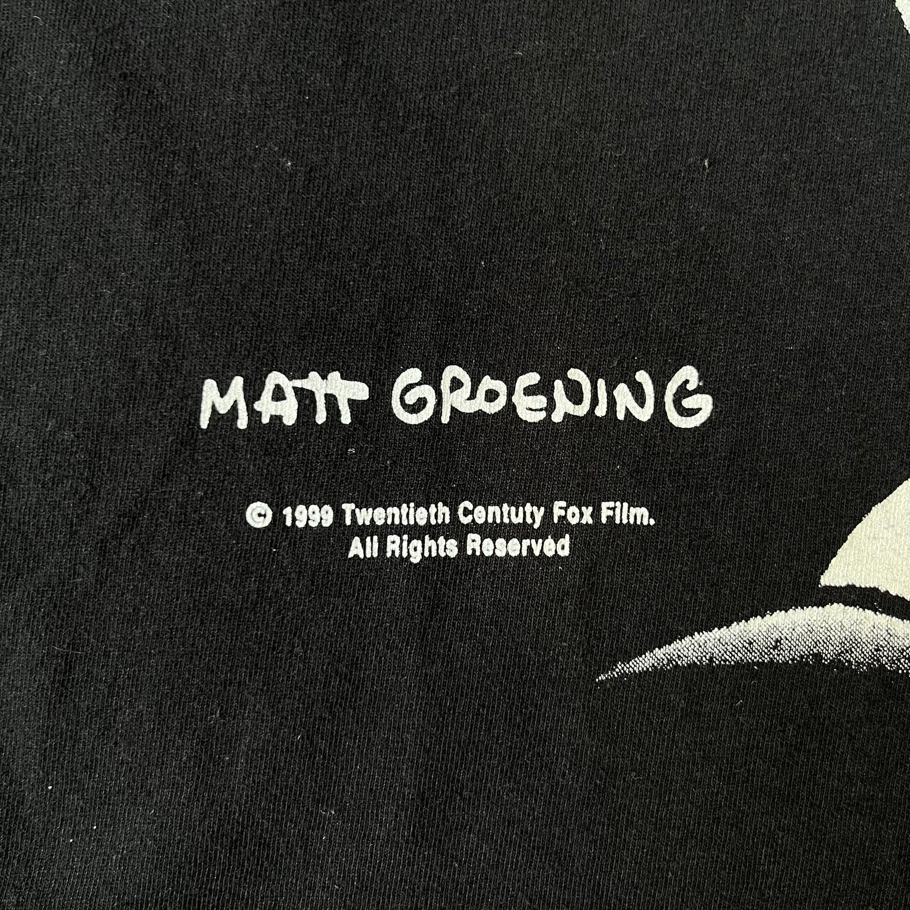 The Simpsons 1999 Bart Face Black Tee features a black T-shirt with a large front graphic of Bart Simpson’s face in black, grey, and white, showing a gritted-teeth expression. Small Matt Groening 1999 licensing text is printed below the graphic.
