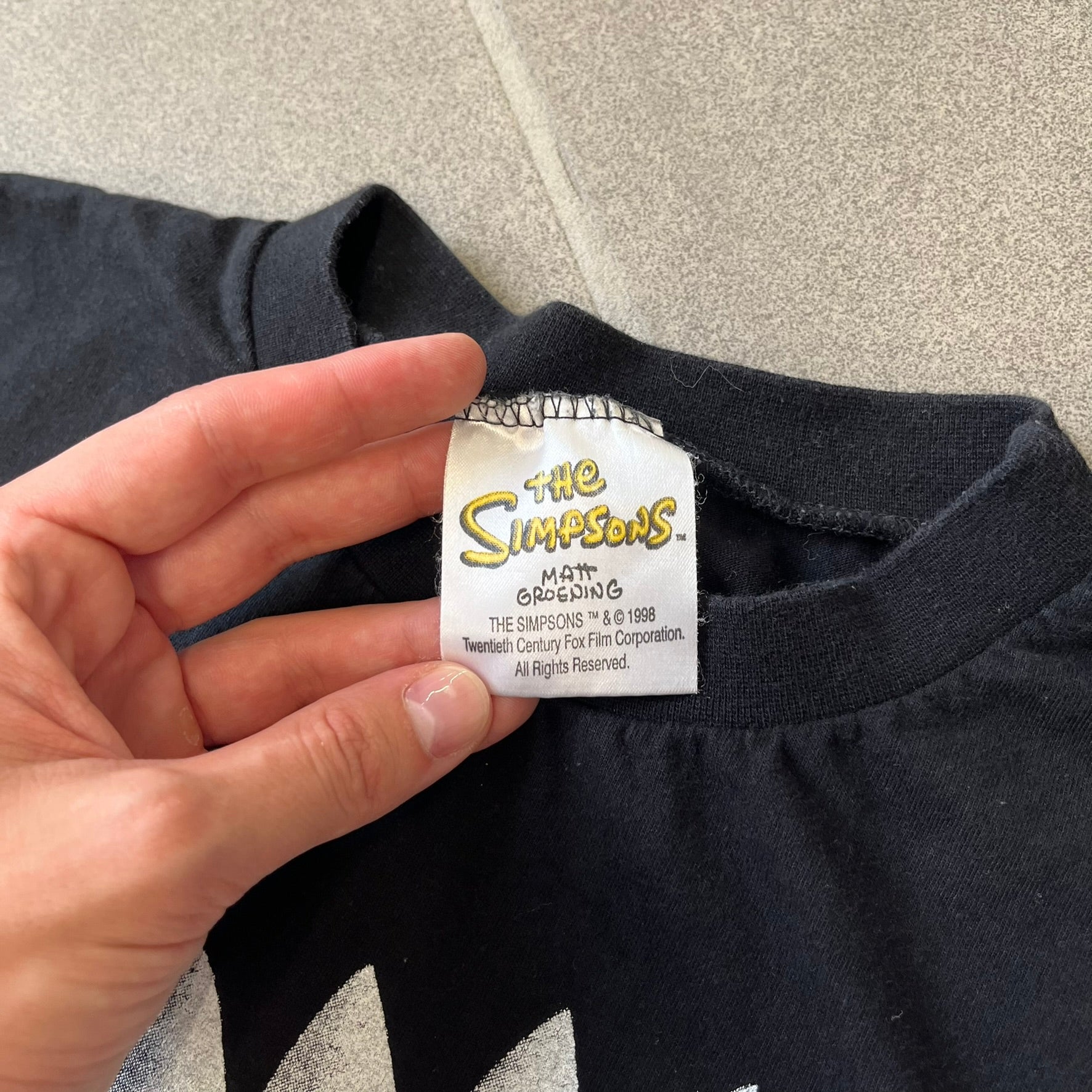 The Simpsons 1999 Bart Face Black Tee features a black T-shirt with a large front graphic of Bart Simpson’s face in black, grey, and white, showing a gritted-teeth expression. Small Matt Groening 1999 licensing text is printed below the graphic.
