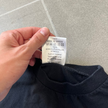 The Simpsons 1999 Bart Face Black Tee features a black T-shirt with a large front graphic of Bart Simpson’s face in black, grey, and white, showing a gritted-teeth expression. Small Matt Groening 1999 licensing text is printed below the graphic.
