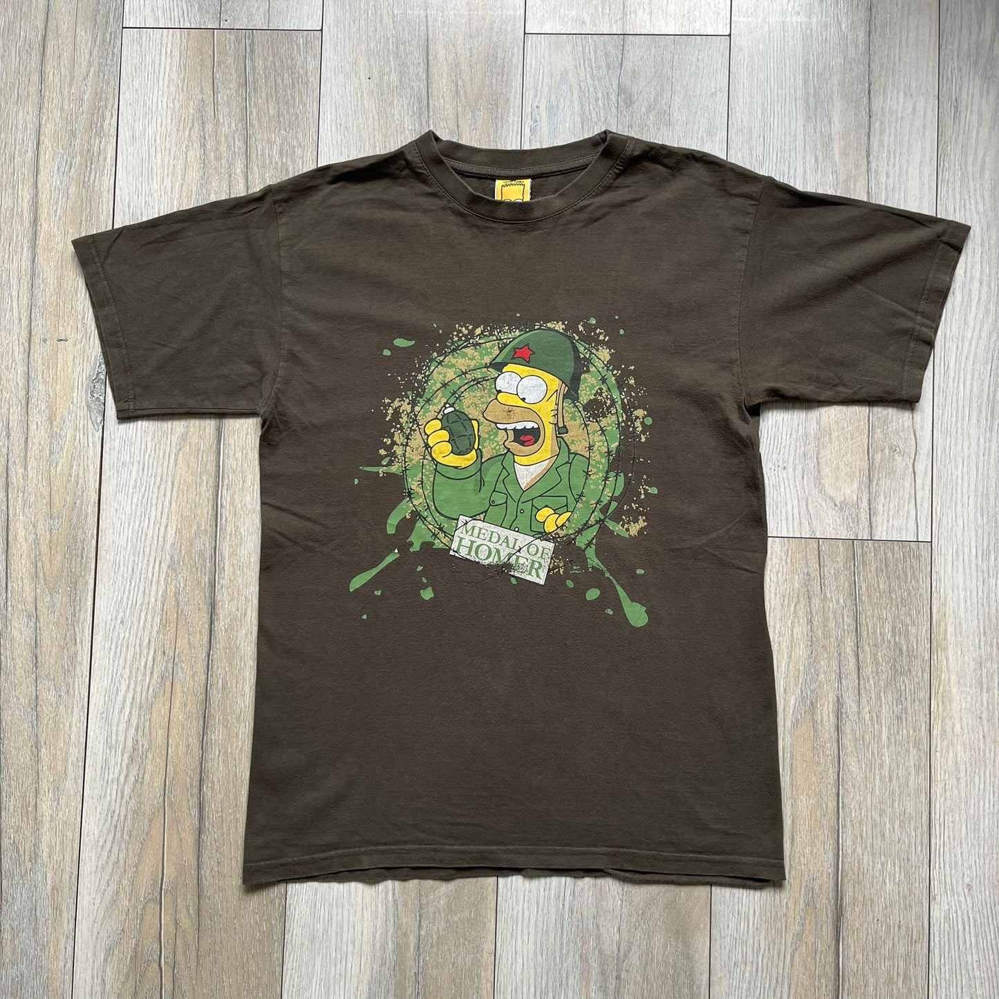 The Simpsons 1999 “Medal of Homer” Tee is a brown T-shirt featuring Homer Simpson in military attire holding a grenade. Below the character is a “Medal of Homer” plaque graphic. The artwork is printed over a green camouflage-style splash background with barbed wire details. The shirt has a yellow tag with Matt Groening’s 1999 licensing and was made in Greece.
