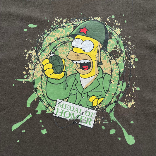 The Simpsons 1999 “Medal of Homer” Tee is a brown T-shirt featuring Homer Simpson in military attire holding a grenade. Below the character is a “Medal of Homer” plaque graphic. The artwork is printed over a green camouflage-style splash background with barbed wire details. The shirt has a yellow tag with Matt Groening’s 1999 licensing and was made in Greece.