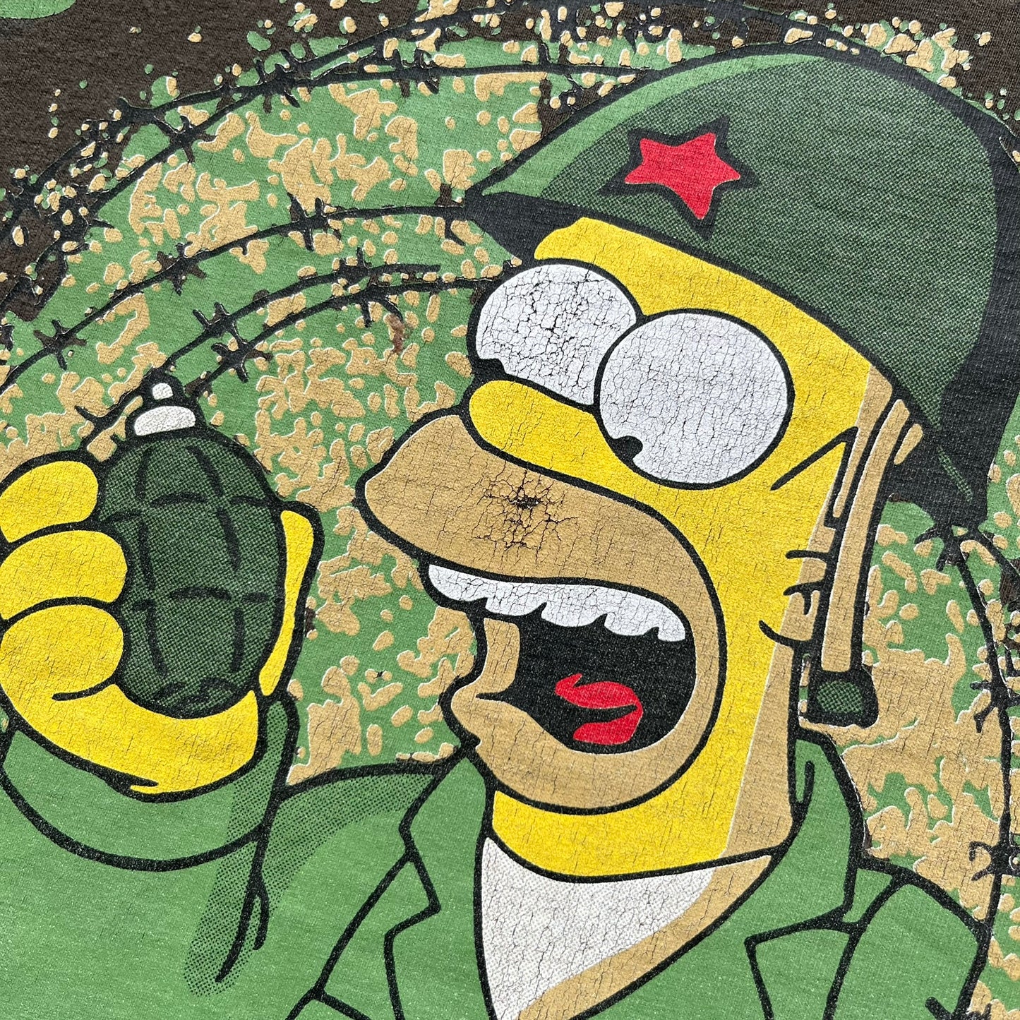 The Simpsons 1999 “Medal of Homer” Tee is a brown T-shirt featuring Homer Simpson in military attire holding a grenade. Below the character is a “Medal of Homer” plaque graphic. The artwork is printed over a green camouflage-style splash background with barbed wire details. The shirt has a yellow tag with Matt Groening’s 1999 licensing and was made in Greece.