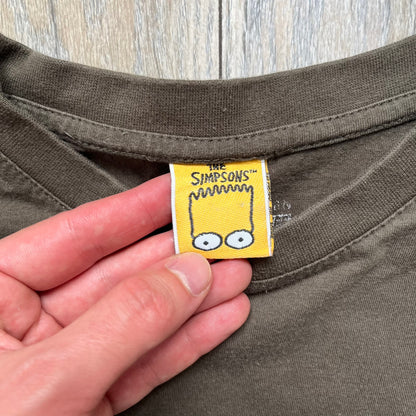 The Simpsons 1999 “Medal of Homer” Tee is a brown T-shirt featuring Homer Simpson in military attire holding a grenade. Below the character is a “Medal of Homer” plaque graphic. The artwork is printed over a green camouflage-style splash background with barbed wire details. The shirt has a yellow tag with Matt Groening’s 1999 licensing and was made in Greece.