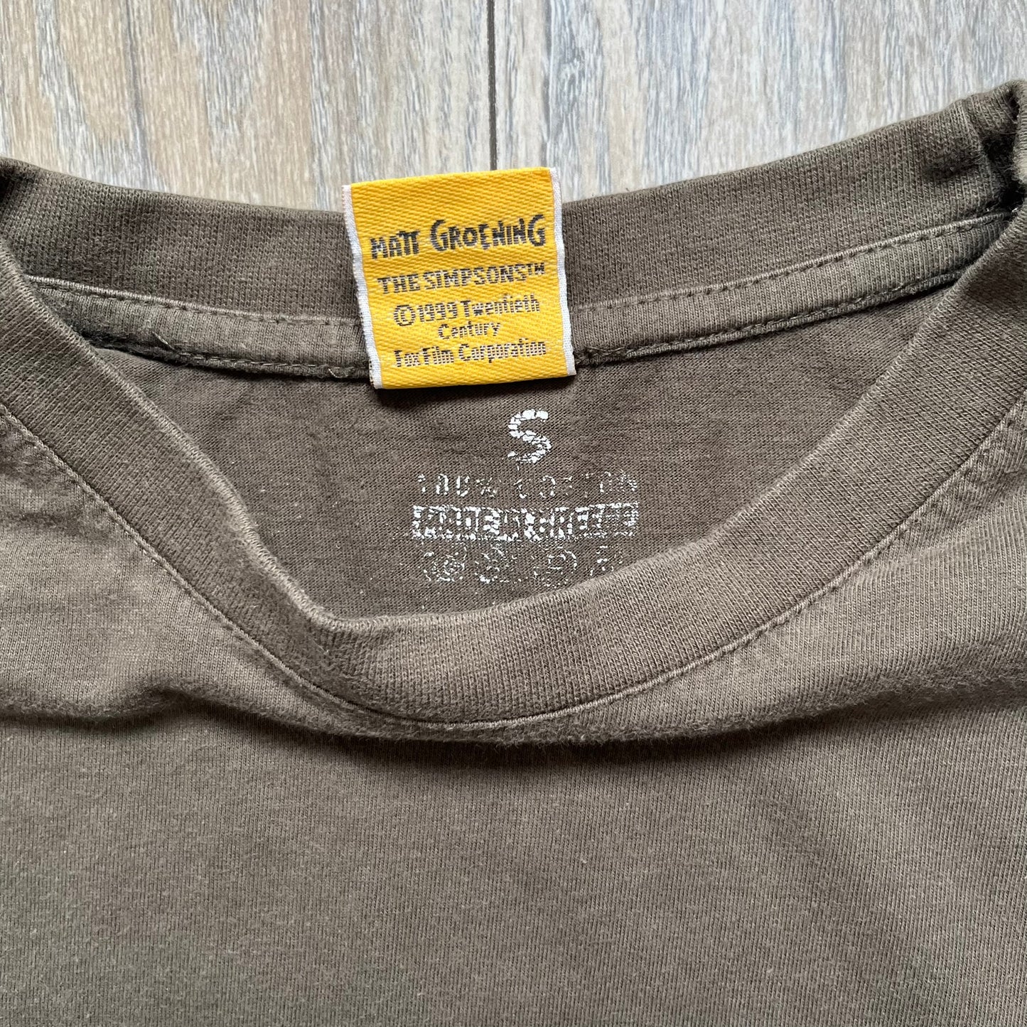 The Simpsons 1999 “Medal of Homer” Tee is a brown T-shirt featuring Homer Simpson in military attire holding a grenade. Below the character is a “Medal of Homer” plaque graphic. The artwork is printed over a green camouflage-style splash background with barbed wire details. The shirt has a yellow tag with Matt Groening’s 1999 licensing and was made in Greece.