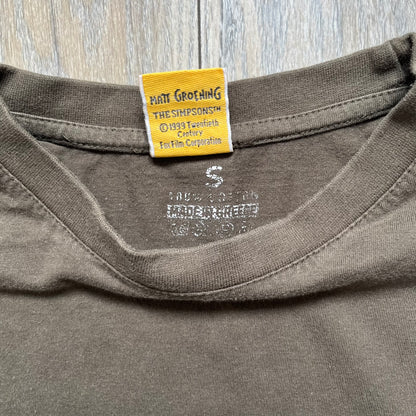 The Simpsons 1999 “Medal of Homer” Tee is a brown T-shirt featuring Homer Simpson in military attire holding a grenade. Below the character is a “Medal of Homer” plaque graphic. The artwork is printed over a green camouflage-style splash background with barbed wire details. The shirt has a yellow tag with Matt Groening’s 1999 licensing and was made in Greece.