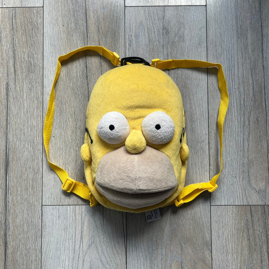 The Simpsons 1990s Homer Simpson Head Backpack is a novelty backpack shaped like a 3D Homer Simpson head. The design is fully yellow with soft sculpted facial features including eyes and nose, a molded mouth, and protruding ears. The hair detail wraps around the back, with the top strands functioning as a hanger. It includes yellow shoulder straps and a single main compartment.