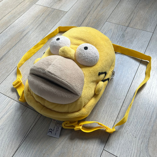 The Simpsons 1990s Homer Simpson Head Backpack is a novelty backpack shaped like a 3D Homer Simpson head. The design is fully yellow with soft sculpted facial features including eyes and nose, a molded mouth, and protruding ears. The hair detail wraps around the back, with the top strands functioning as a hanger. It includes yellow shoulder straps and a single main compartment.