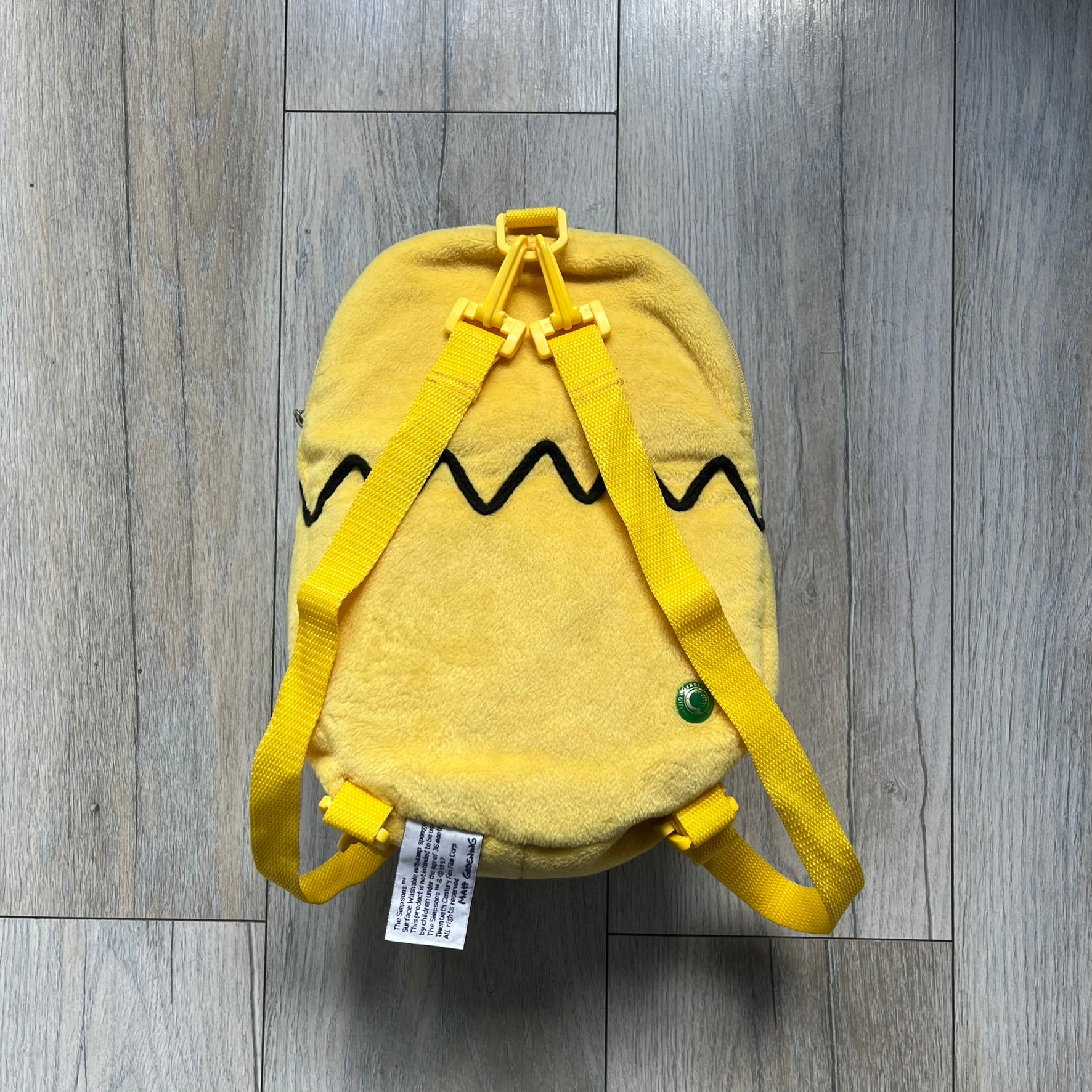 The Simpsons 1990s Homer Simpson Head Backpack is a novelty backpack shaped like a 3D Homer Simpson head. The design is fully yellow with soft sculpted facial features including eyes and nose, a molded mouth, and protruding ears. The hair detail wraps around the back, with the top strands functioning as a hanger. It includes yellow shoulder straps and a single main compartment.