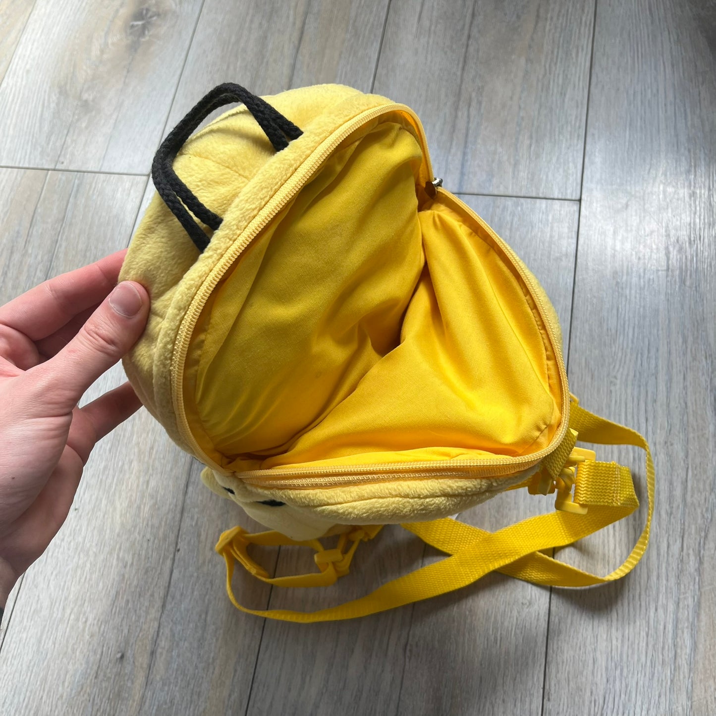 The Simpsons 1990s Homer Simpson Head Backpack is a novelty backpack shaped like a 3D Homer Simpson head. The design is fully yellow with soft sculpted facial features including eyes and nose, a molded mouth, and protruding ears. The hair detail wraps around the back, with the top strands functioning as a hanger. It includes yellow shoulder straps and a single main compartment.