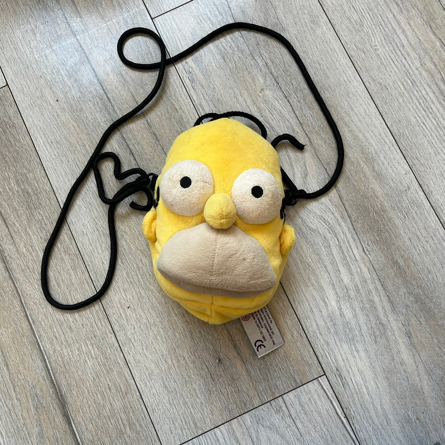 The Simpsons 1990s Homer Simpson Head Shoulder bag is shaped like a 3D Homer Simpson head. The design is yellow with soft sculpted facial features including eyes and nose, a molded beige mouth, and protruding ears. The hair detail wraps around the back, with the top strands functioning as a hanger. It includes a black string and a single main compartment.