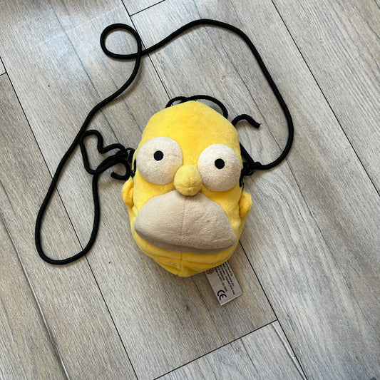 The Simpsons 1990s Homer Simpson Head Shoulder bag is shaped like a 3D Homer Simpson head. The design is yellow with soft sculpted facial features including eyes and nose, a molded beige mouth, and protruding ears. The hair detail wraps around the back, with the top strands functioning as a hanger. It includes a black string and a single main compartment.