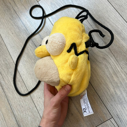 The Simpsons 1990s Homer Simpson Head Shoulder bag is shaped like a 3D Homer Simpson head. The design is yellow with soft sculpted facial features including eyes and nose, a molded beige mouth, and protruding ears. The hair detail wraps around the back, with the top strands functioning as a hanger. It includes a black string and a single main compartment.