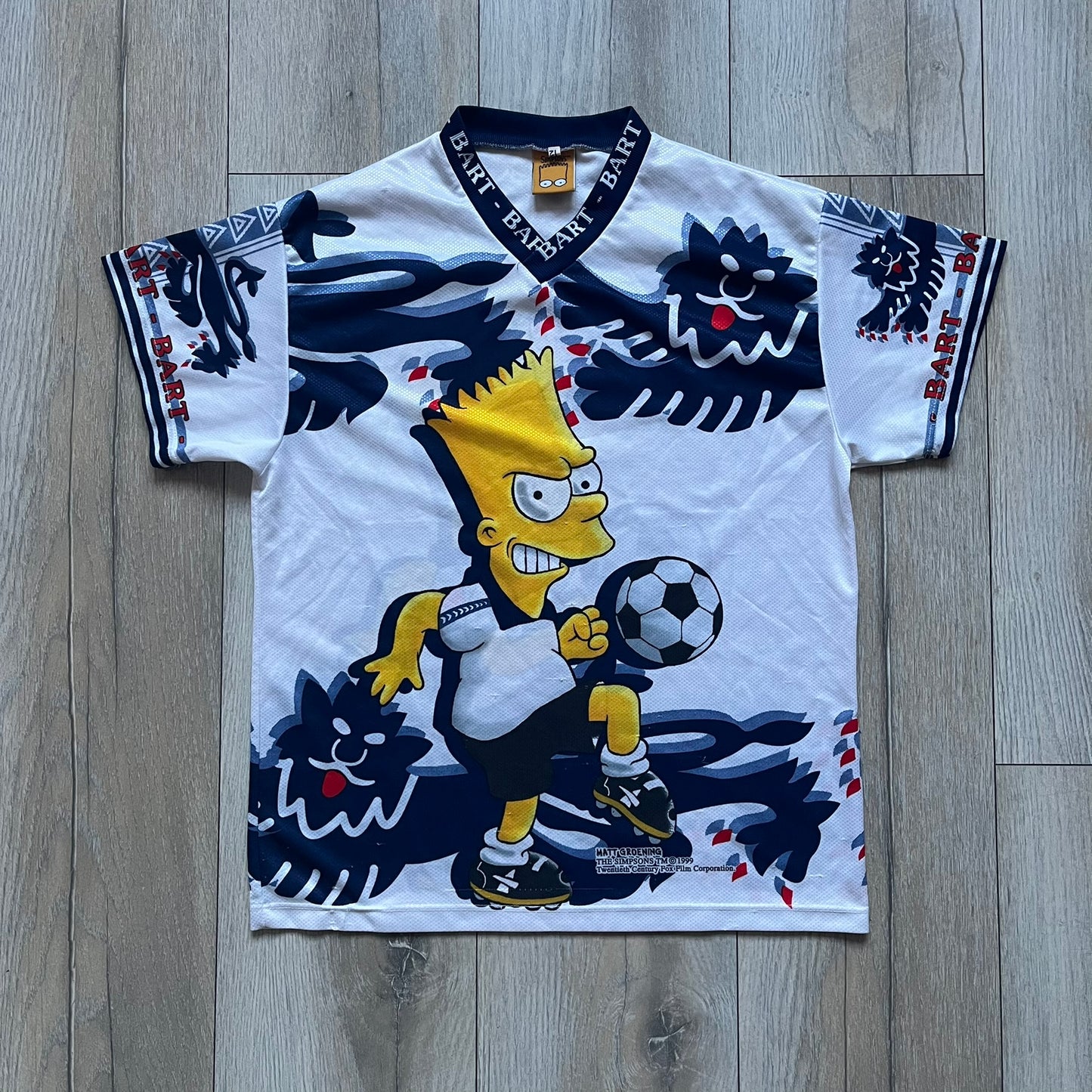 Officially licensed 1999 The Simpsons World Cup T-shirt for England. Lightweight white polyester tee featuring an all-over graphic design. The front shows Bart Simpson kicking a football, dressed in a white tee, with multiple England three-lions emblems layered across the background from top to bottom. Red “Bart” text details appear on the sleeve hems, with Matt Groening / The Simpsons ©1999 licensing printed beneath the main graphic.&nbsp;The back mirrors the artwork in a flipped layout, showing Bart from 