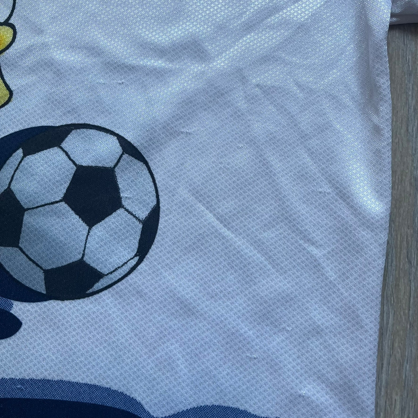 Officially licensed 1999 The Simpsons World Cup T-shirt for England. Lightweight white polyester tee featuring an all-over graphic design. The front shows Bart Simpson kicking a football, dressed in a white tee, with multiple England three-lions emblems layered across the background from top to bottom. Red “Bart” text details appear on the sleeve hems, with Matt Groening / The Simpsons ©1999 licensing printed beneath the main graphic.&nbsp;The back mirrors the artwork in a flipped layout, showing Bart from 