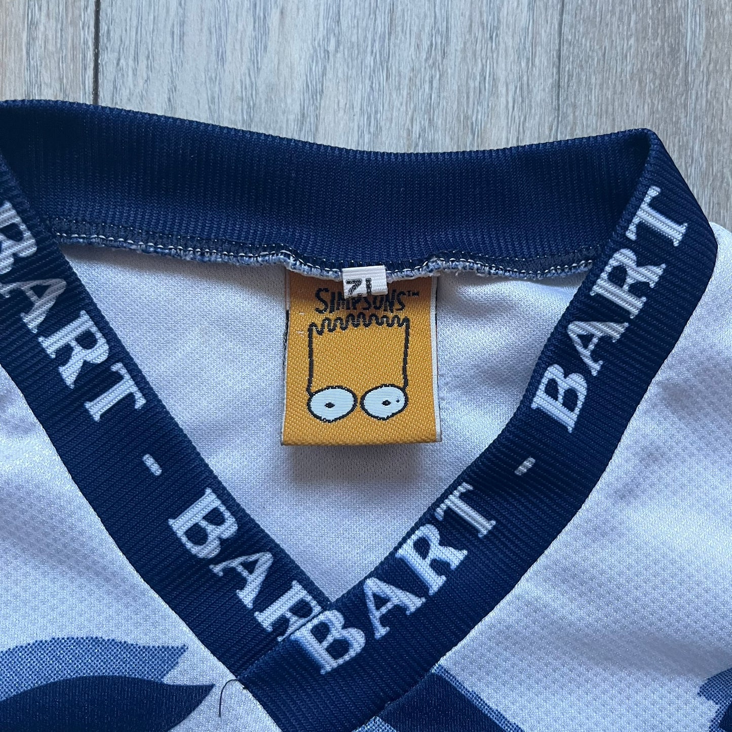 Officially licensed 1999 The Simpsons World Cup T-shirt for England. Lightweight white polyester tee featuring an all-over graphic design. The front shows Bart Simpson kicking a football, dressed in a white tee, with multiple England three-lions emblems layered across the background from top to bottom. Red “Bart” text details appear on the sleeve hems, with Matt Groening / The Simpsons ©1999 licensing printed beneath the main graphic.&nbsp;The back mirrors the artwork in a flipped layout, showing Bart from 