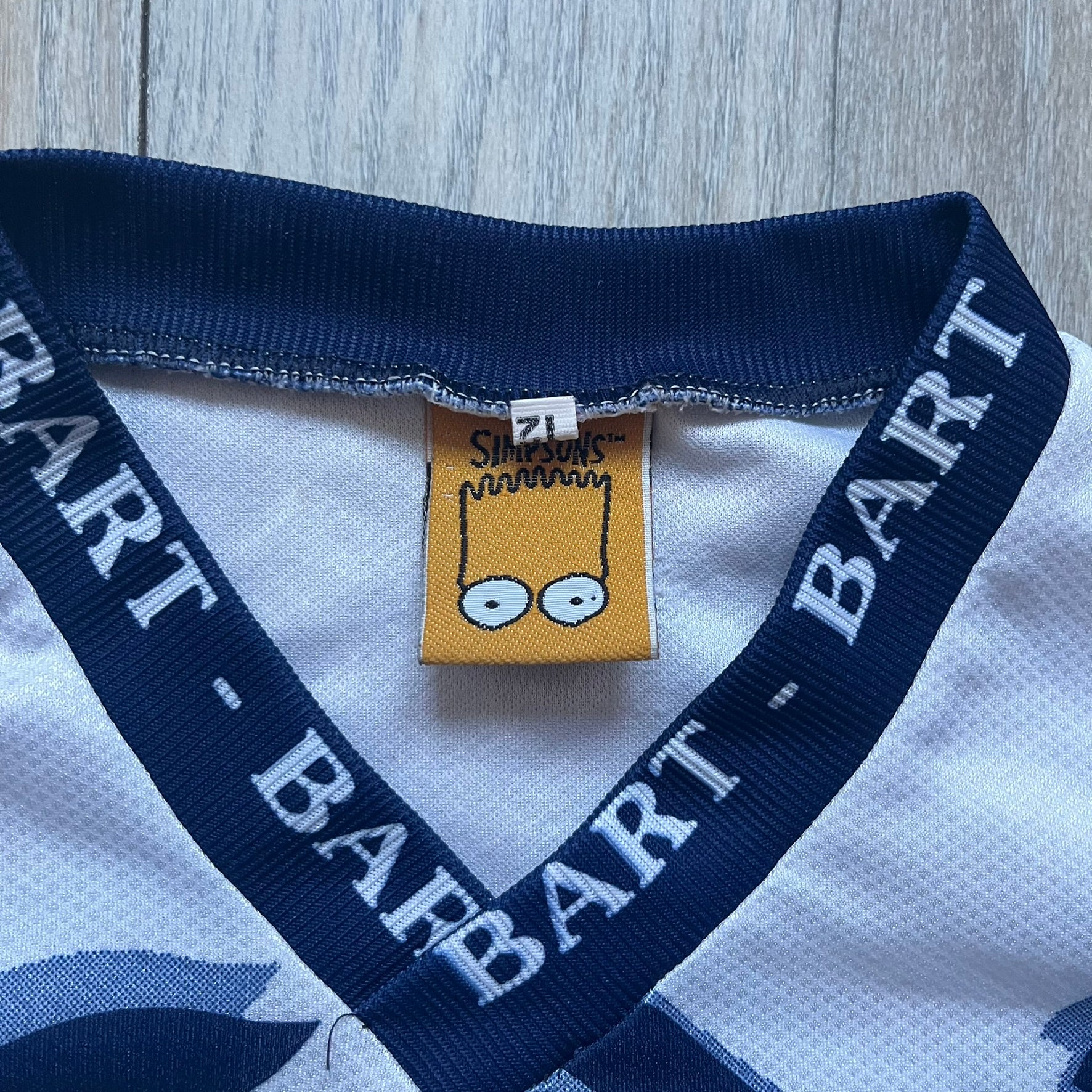 Officially licensed 1999 The Simpsons World Cup T-shirt for England. Lightweight white polyester tee featuring an all-over graphic design. The front shows Bart Simpson kicking a football, dressed in a white tee, with multiple England three-lions emblems layered across the background from top to bottom. Red “Bart” text details appear on the sleeve hems, with Matt Groening / The Simpsons ©1999 licensing printed beneath the main graphic.&nbsp;The back mirrors the artwork in a flipped layout, showing Bart from 